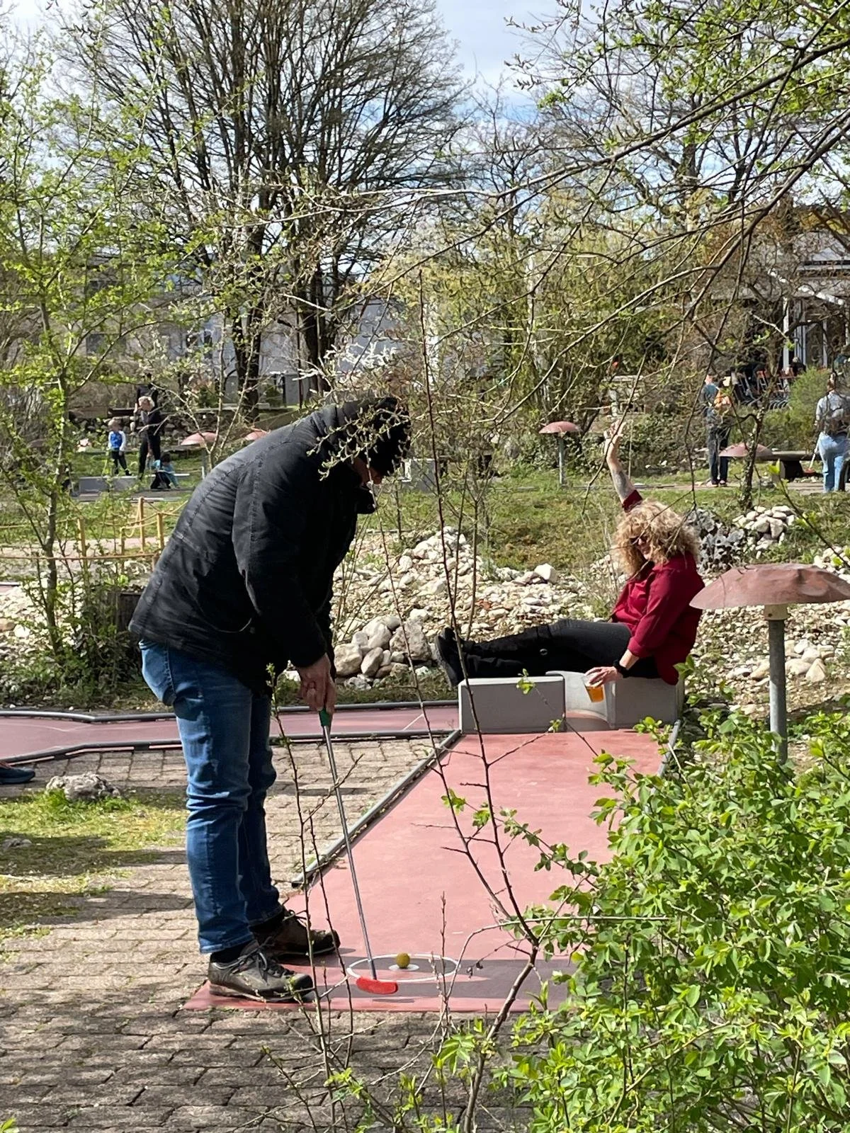 Zwei Menschen spielen Minigolf im Park, einer holt den Golfball vom Loch, die andere sitzt auf einer Bank mit einem pinken Regenschirm. Es sind Bäume und andere Personen im Hintergrund.