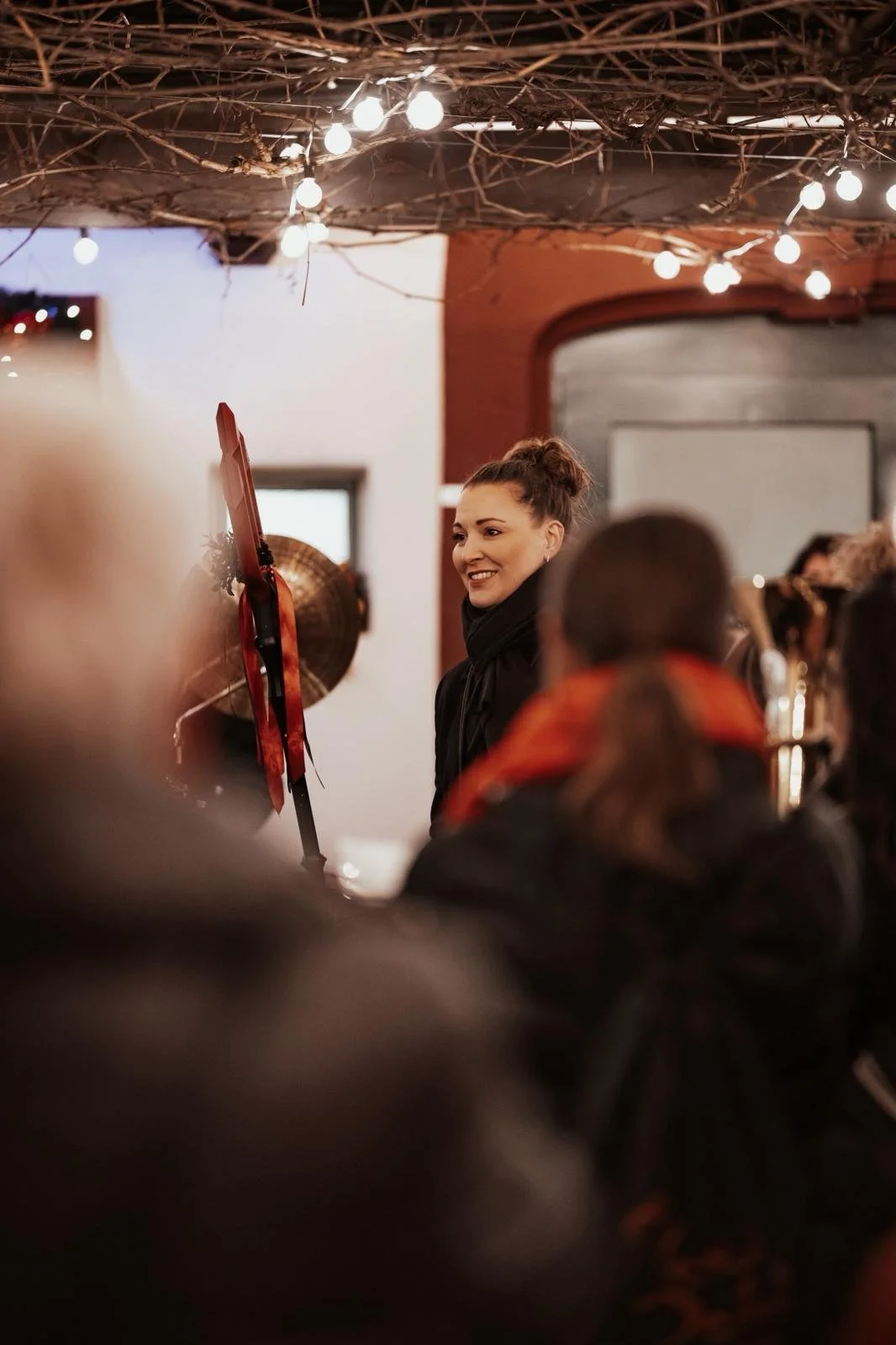 Eine junge Frau lächelt während einer Veranstaltung, umgeben von mehreren Menschen, unter einer Dekoration aus Zweigen und Lichterketten im Innenraum.