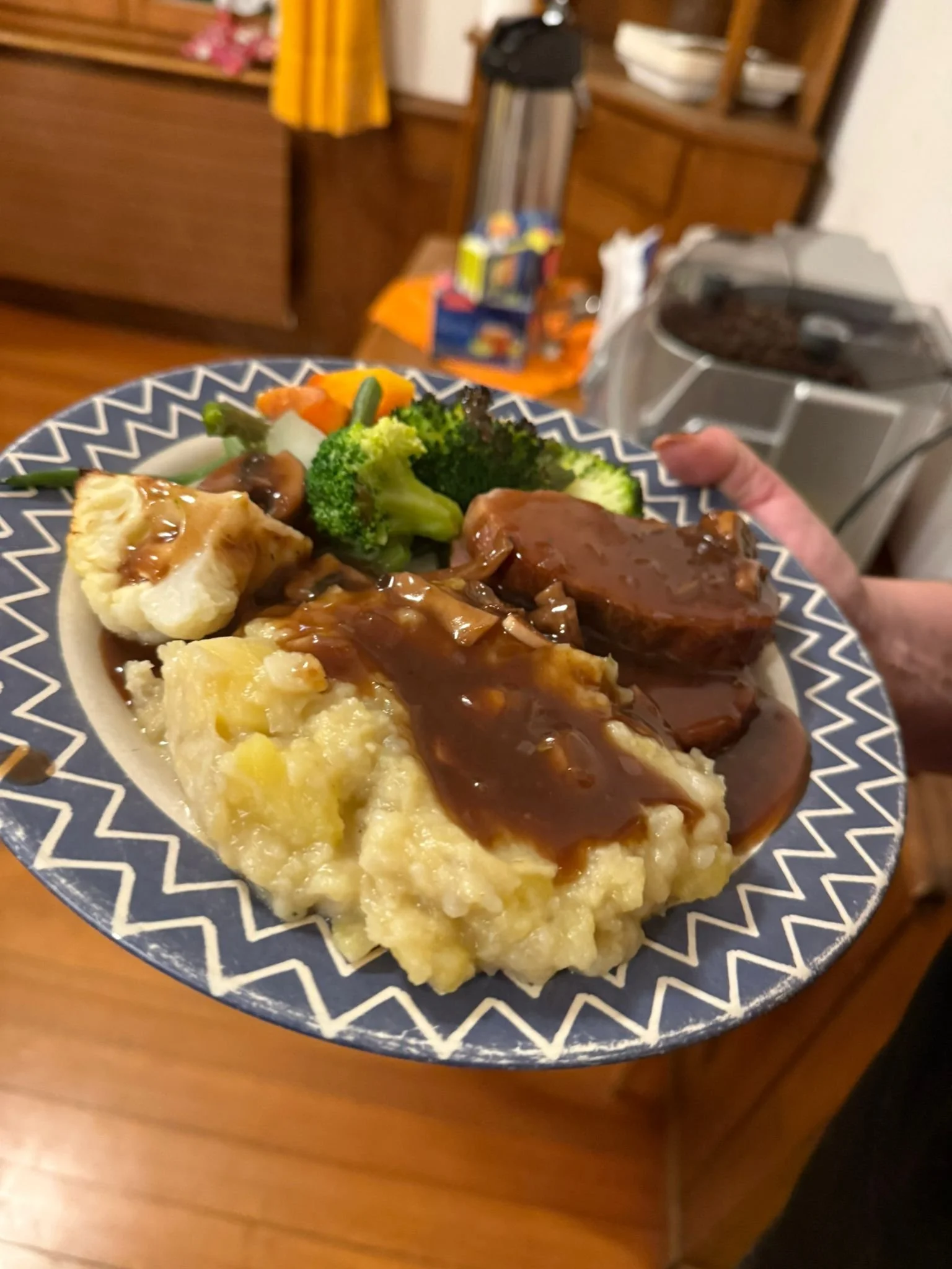 Ein Teller mit Hackbraten, Kartoffelbrei mit brauner Soße, Brokkoli, Blumenkohl und gemischtem Gemüse auf einem blauen und weißen Teller. Im Hintergrund ein Raum mit Holzmöbeln und einem Getränkespender.