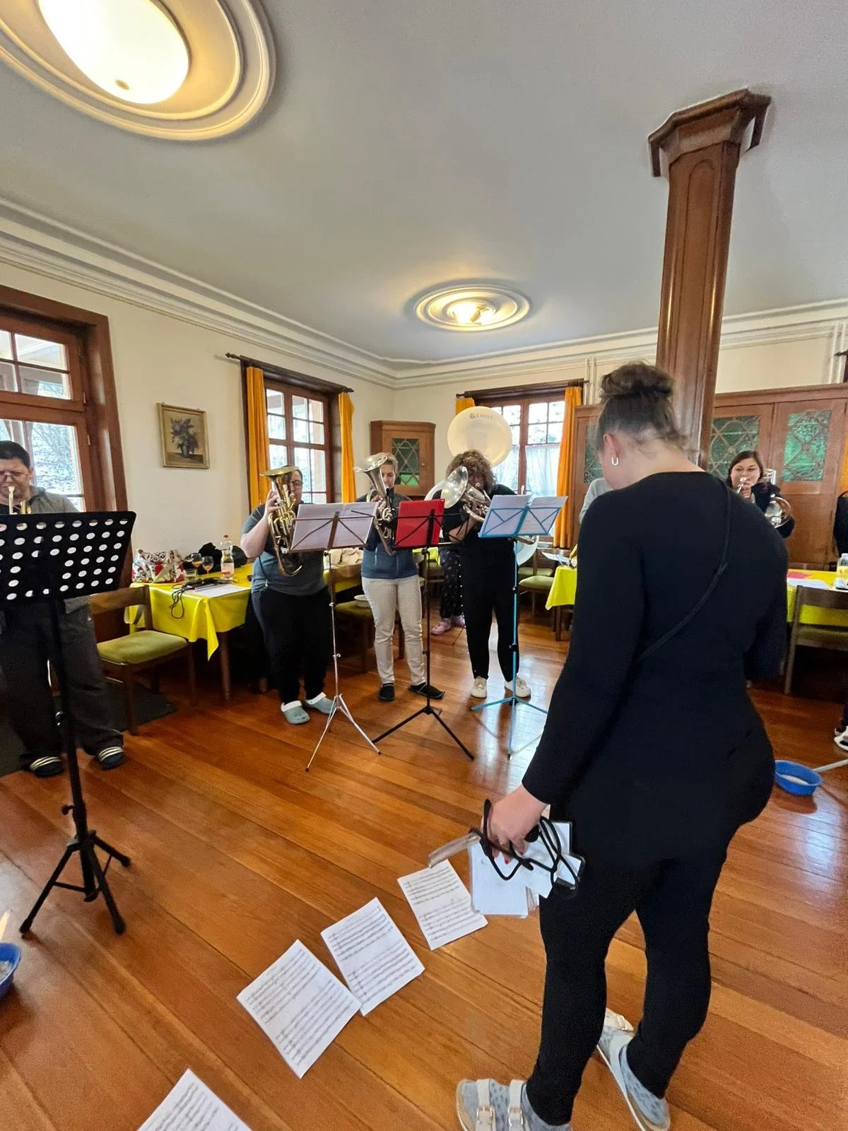 Eine Gruppe von Musikern, die in einem Raum mit holzvertäfelten Wänden und großen Fenstern ein Proben- oder Konzert vorbereitet. Im Vordergrund steht eine Frau in schwarzer Kleidung, die Noten in der Hand hält.