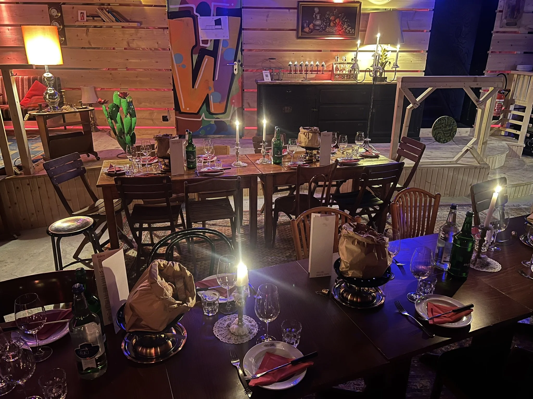 Gemütlich gedeckter Esstisch mit Kerzen, Gläsern, Flaschen und Besteck in einem festlich gestalteten Raum, im Hintergrund bunte Wandgestaltung und Dekorationen.