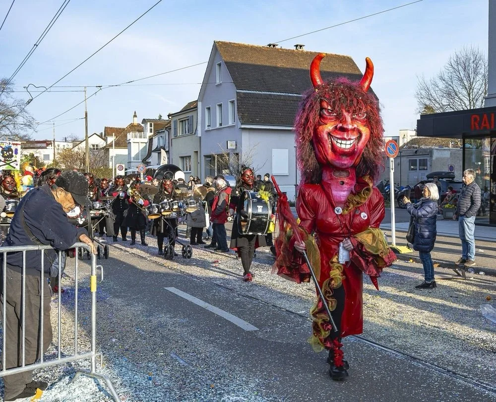 Karnevals- oder Fasnachtsumzug mit einer großen Teufelsmaske in rotem Outfit und Maskierte im Hintergrund, Straßenumzug in einer Stadt, sonniger Tag.