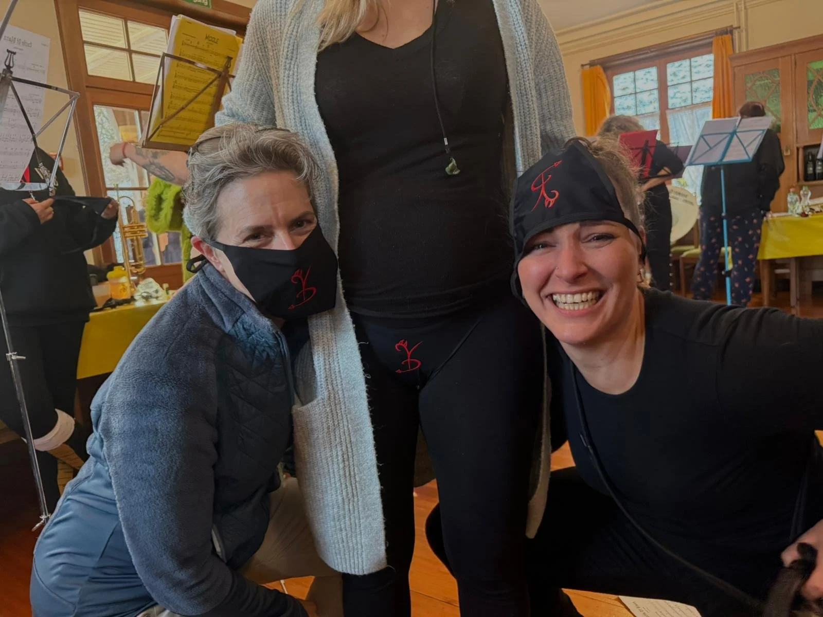 Drei Frauen in einem Raum, feiern und lachen. Zwei Frauen tragen schwarze Masken mit rotem Logo, eine Frau im Hintergrund trägt eine Bandana. Der Raum hat Holzwände und große Fenster mit gelben Vorhängen, Tische mit gelbem Tischtuch, Musiker im Hinte