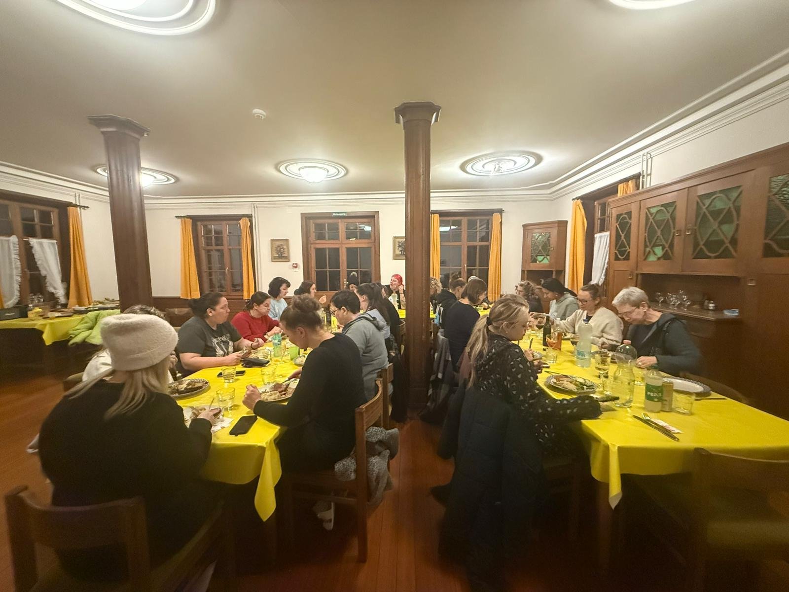 Menschen sitzen an zwei Tischen in einem großen Speiseraum, der mit holzverkleideten Wänden und gelben Tischdecken gestaltet ist, beim Abendessen.