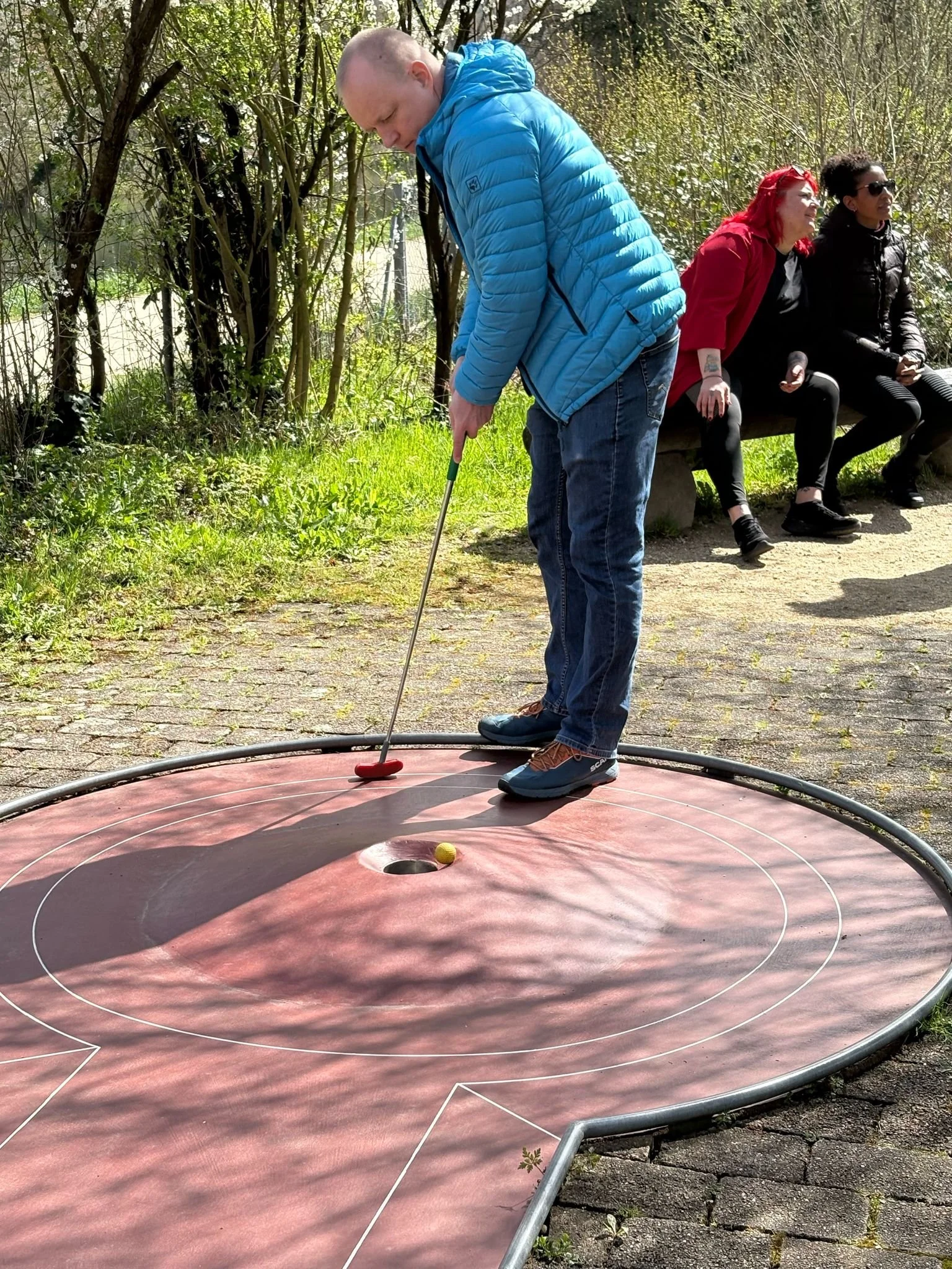 Ein Mann spielt Mini-Golf auf einer kleinen Outdoor-Anlage, während zwei Frauen auf einer Bank im Hintergrund sitzen und sich unterhalten, umgeben von Bäumen und Grün.