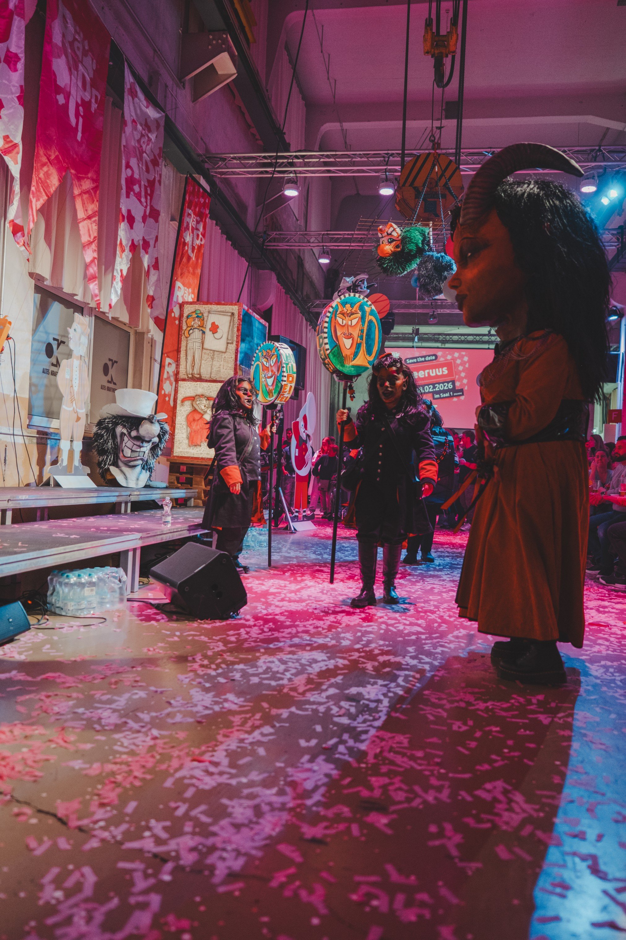 Menschen in Karnevals- oder Faschingskostümen auf einer Party, mit bunten Dekorationen und confetti auf dem Boden, in einer festlich beleuchteten Halle.