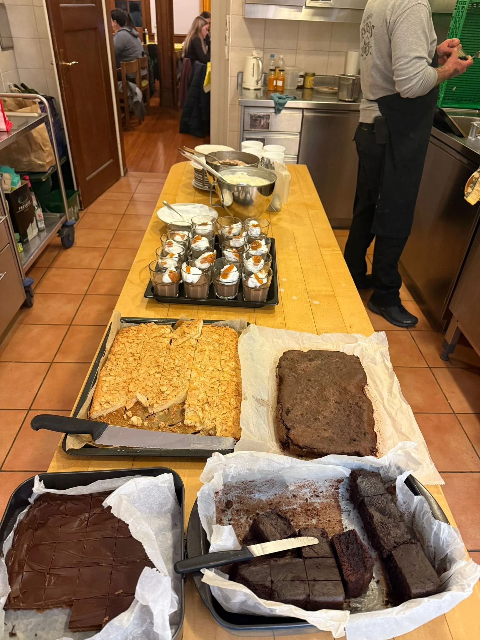 Auf einem Holz Tisch stehen verschiedene Kuchen und Desserts, darunter Schokoladenkuchen, Apfelkuchen, Tiramisu und andere Leckereien, während im Hintergrund Menschen in einer Restaurantküche an Tischen sitzen.