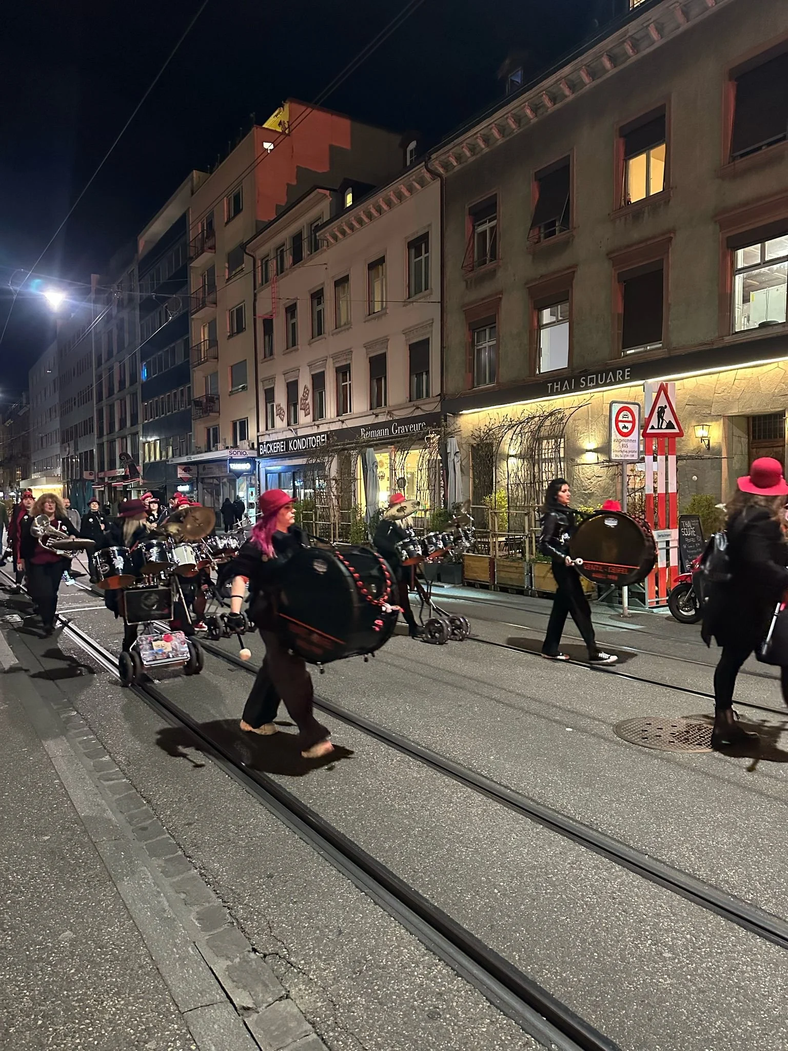 Menschen ziehen in einer Parade mit Musikinstrumenten durch eine Stadtstraße bei Nacht, im Hintergrund mehrstöckige Gebäude und Lichter.