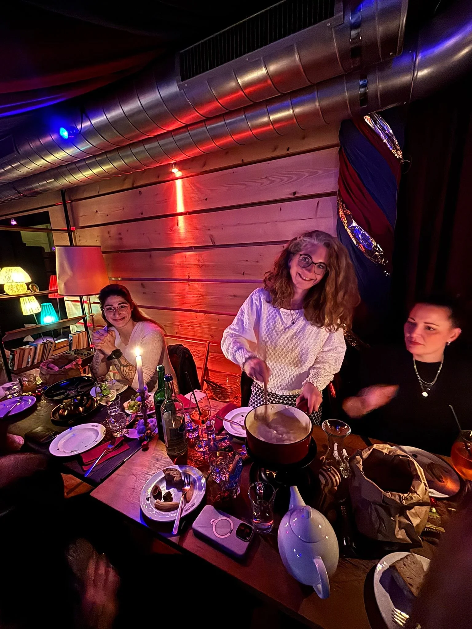 Drei Frauen sitzen an einem festlich gedeckten Tisch mit Speisen und Getränken bei einer Feier, eine Frau in der Mitte steht mit einem Topf in der Hand und lacht, die Umgebung ist warm beleuchtet mit bunten Lampen.