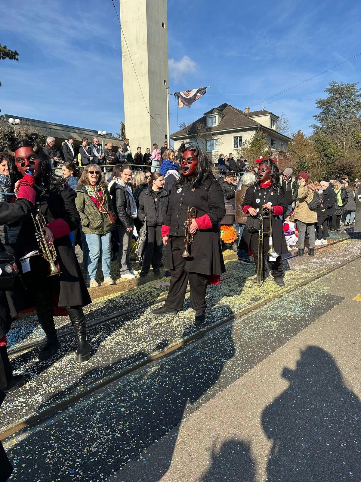 Menschen in Karnevalskostümen mit Dämonenmasken, die auf einem Umzug mit Konfetti auf der Straße stehen, während Zuschauer im Hintergrund zuschauen.
