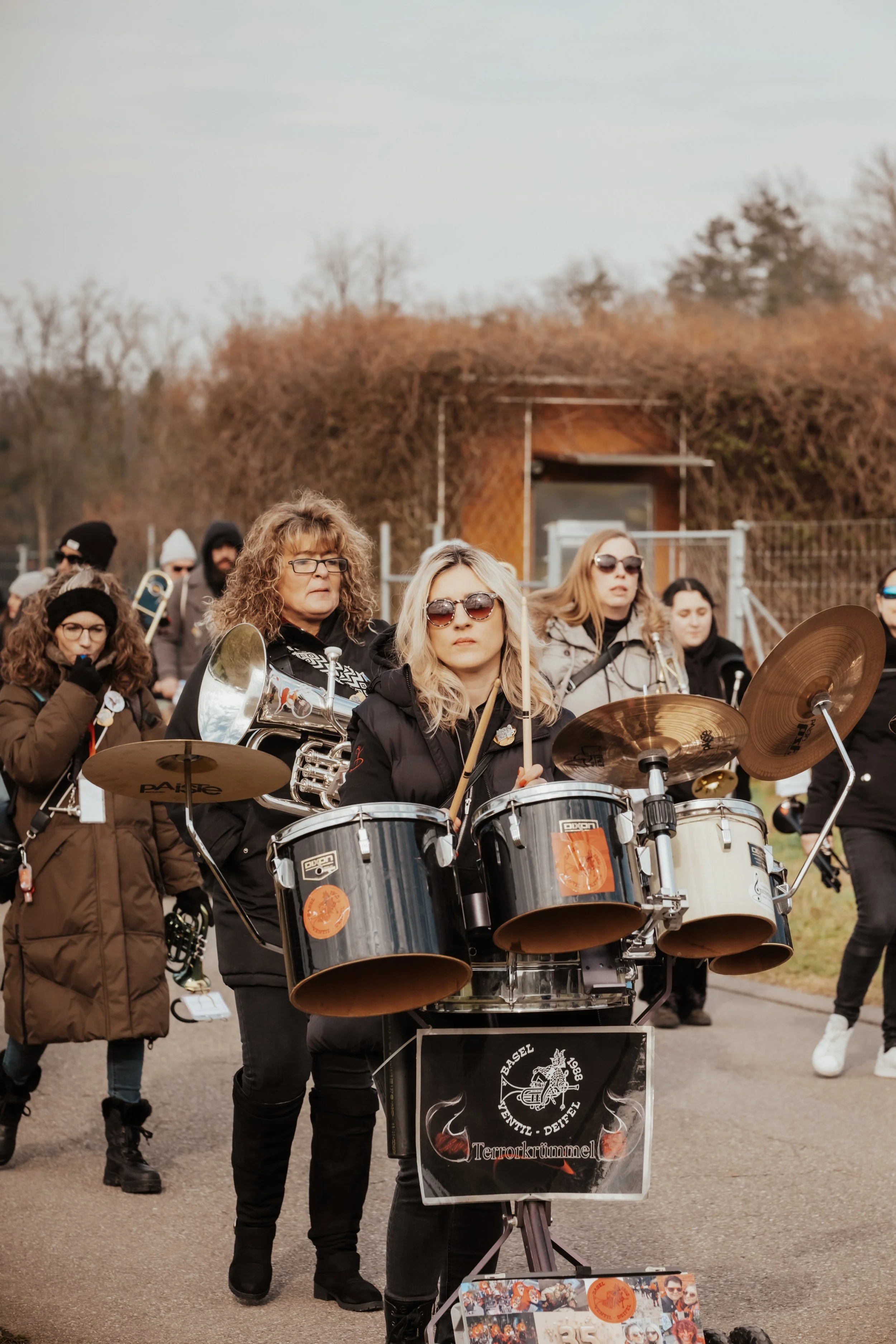 Eine Gruppe von Menschen, darunter eine Frau, die Schlagzeug spielt, bei einer outdoor Parade oder Veranstaltung.