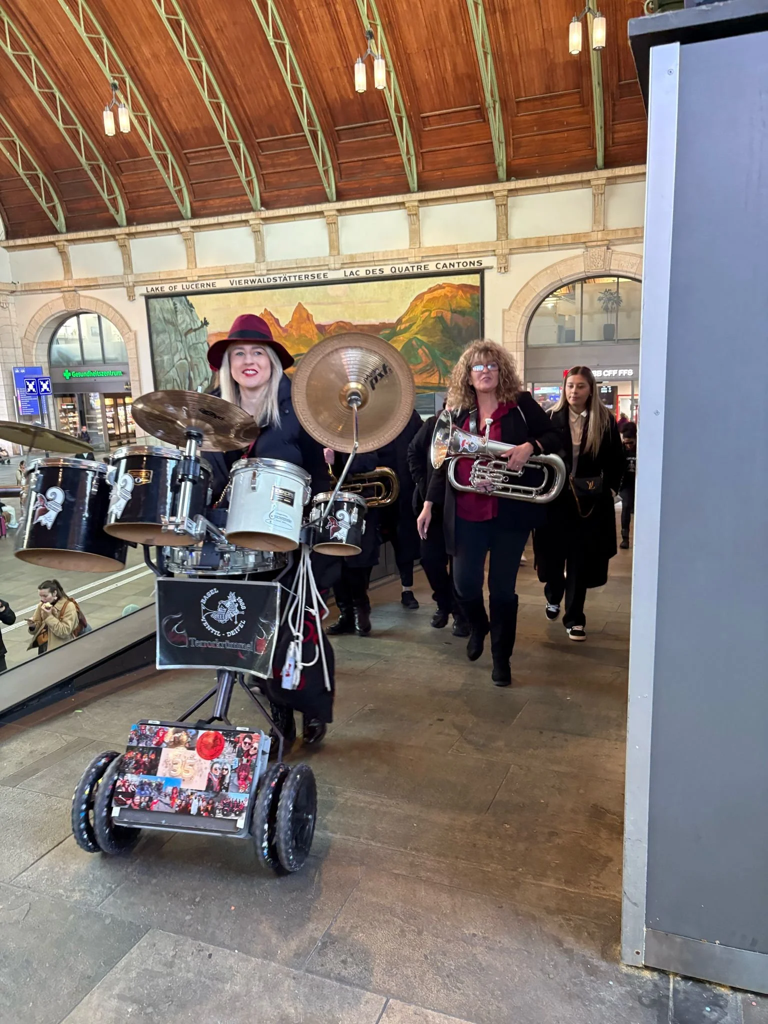 Musikerinnen mit verschiedenen Instrumenten, darunter Schlagzeug, Klarinette und Eufonium, laufen durch einen Bahnhofshalle.