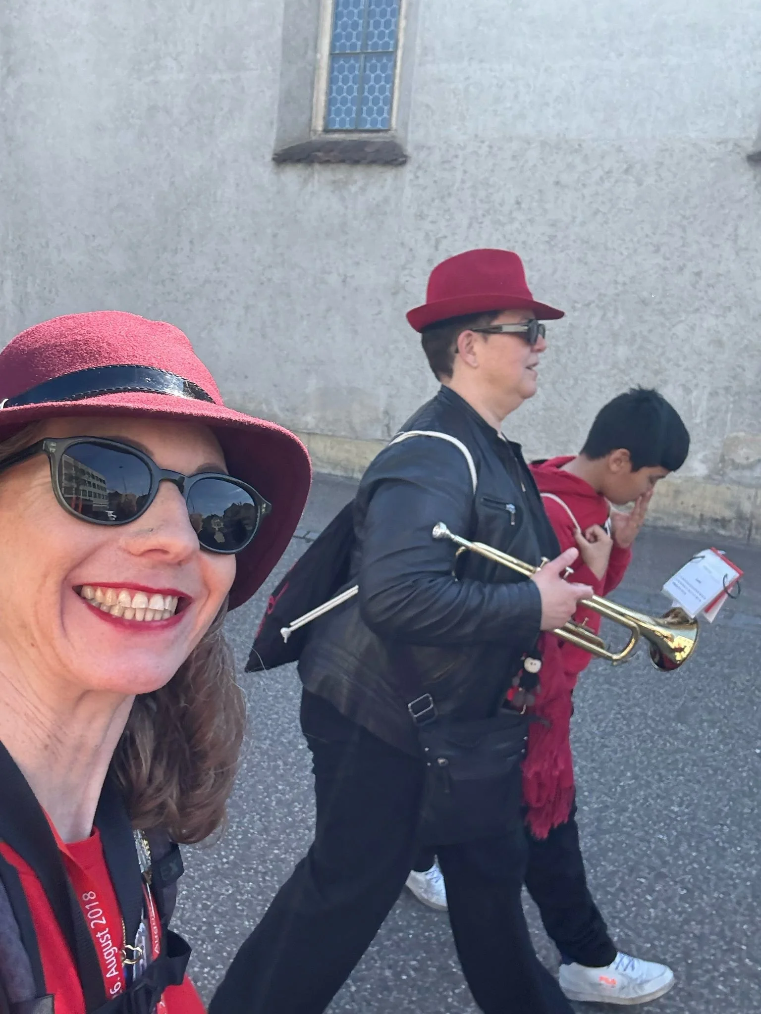 Drei Menschen gehen spazieren, eine Frau im Vordergrund trägt einen roten Hut und Sonnenbrille, im Hintergrund eine Frau mit rotem Hut, die eine Trompete hält, und ein Kind in roter Jacke.