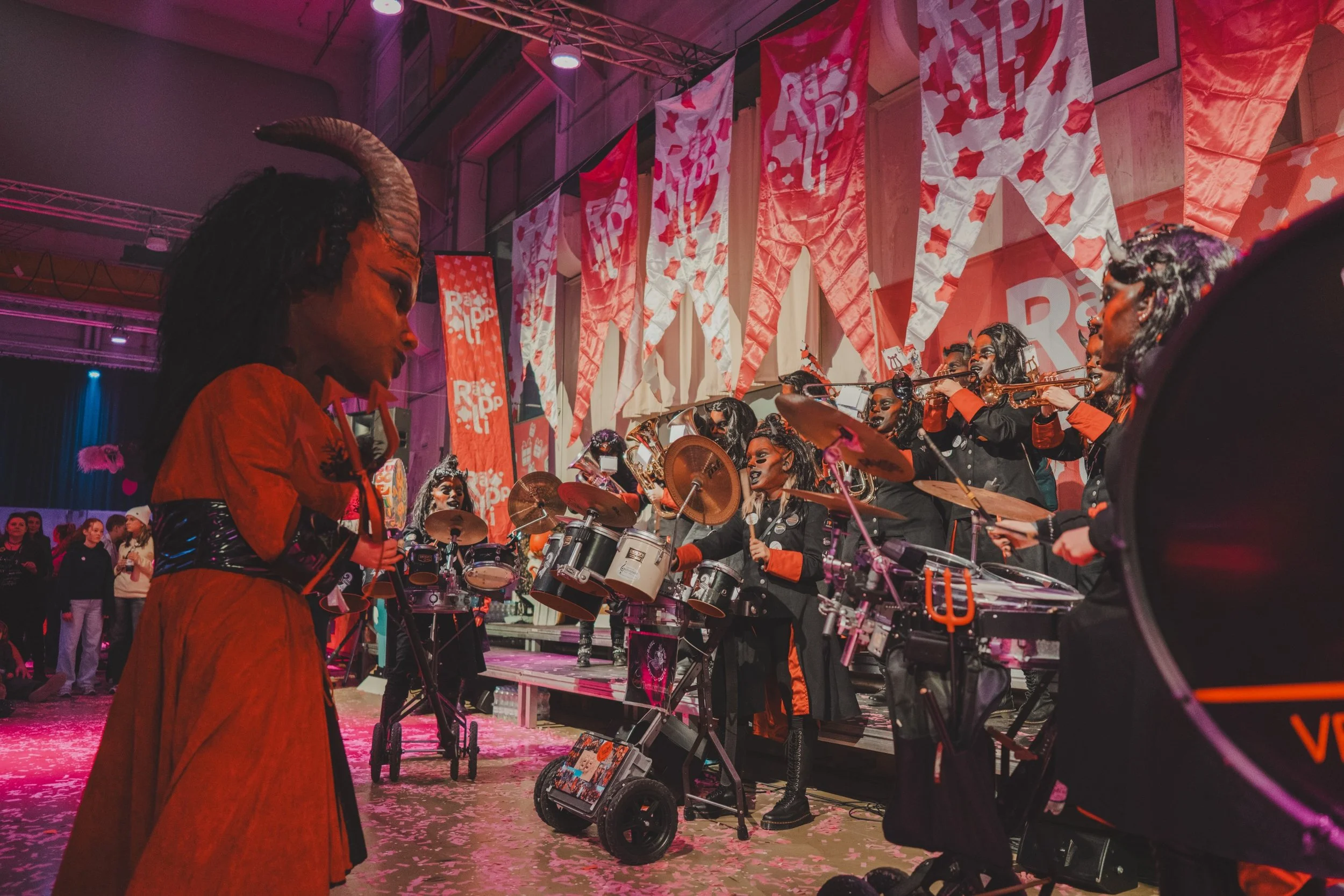 Eine Gruppe von Musikerinnen in Kostümen mit schwarzen Locken und Gesichtsbügel während einer Aufführung auf einer Bühne, umgeben von rot-weiß dekorierten Banner und Flaggen.