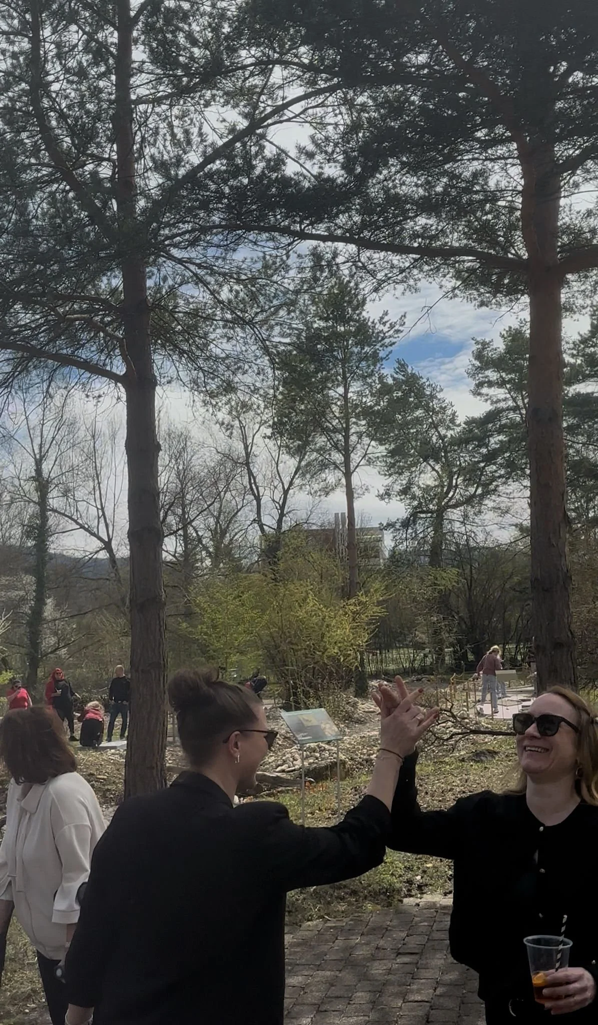 Zwei Frauen high-fiven sich lachend im Freien, im Hintergrund sind weitere Personen und Bäume.
