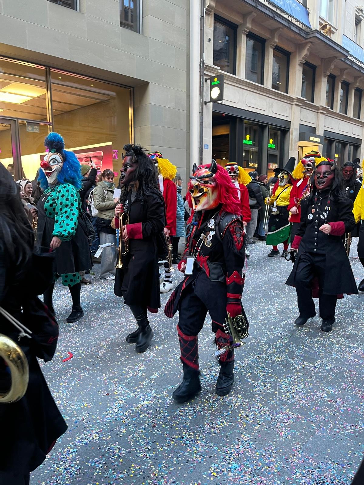 Menschen in farbenfrohen Clown- und Masken-Kostümen, die an einem Straßenumzug teilnehmen, mit Konfetti auf dem Boden.