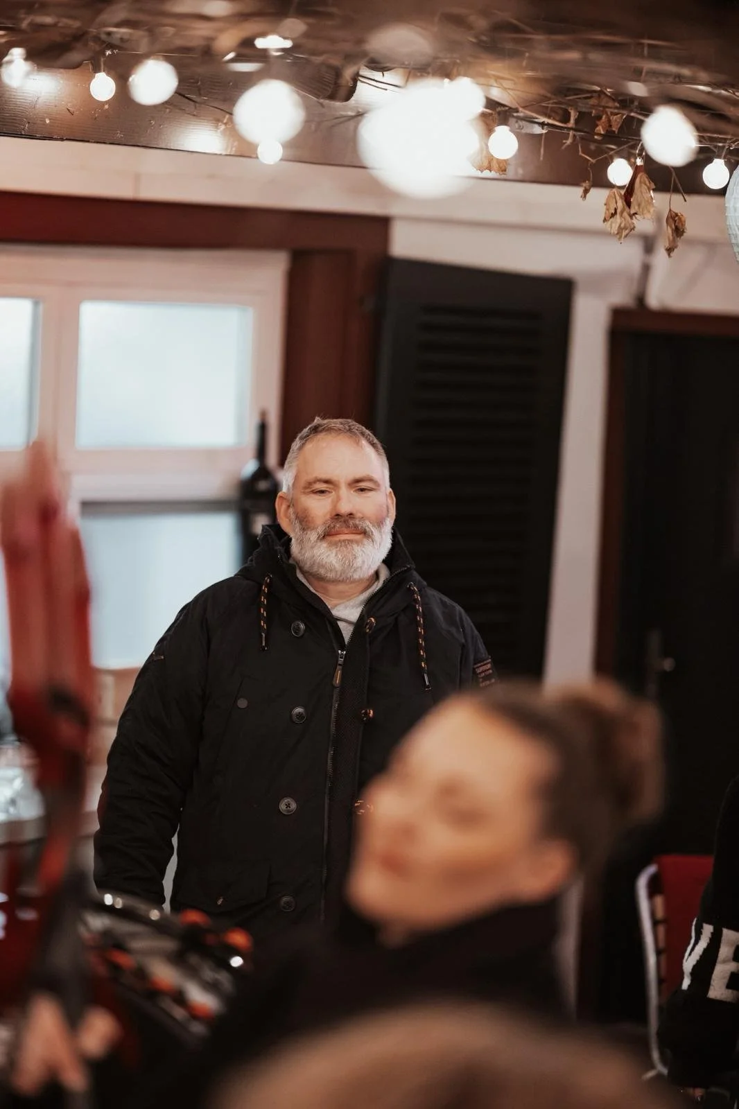 Ein Mann mit grauem Bart und schwarzen Jacke steht in einem Raum mit gestrichenen Wänden und Deko-Lichtern an der Decke, während eine Person im Vordergrund unscharf ist.