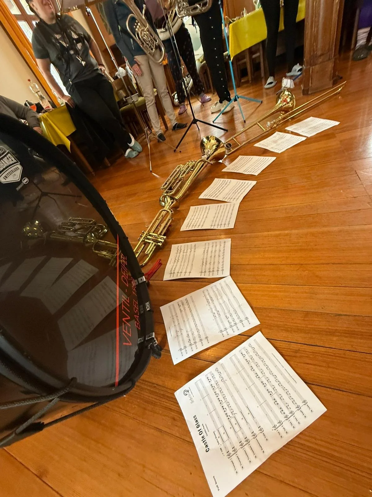 Musikinstrumente und Notenblätter auf Holztisch bei einer musikalischen Darbietung in einem Raum mit Menschen im Hintergrund.