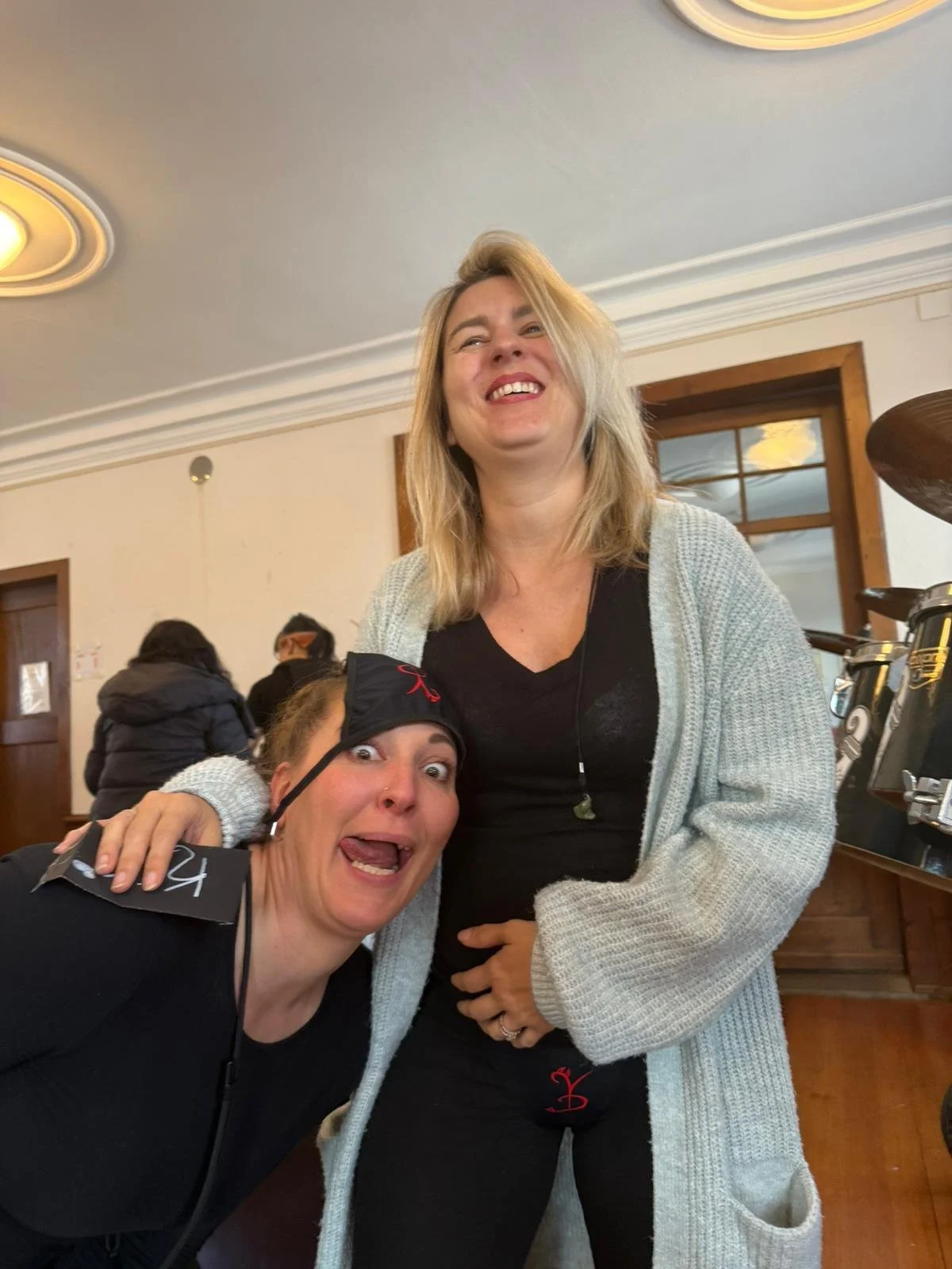 Zwei lachende Frauen, eine steht hinter der anderen, die kniet. Sie erscheinen fröhlich und haben Spaß. Im Hintergrund sieht man andere Menschen und einen Raum mit Holztüren.