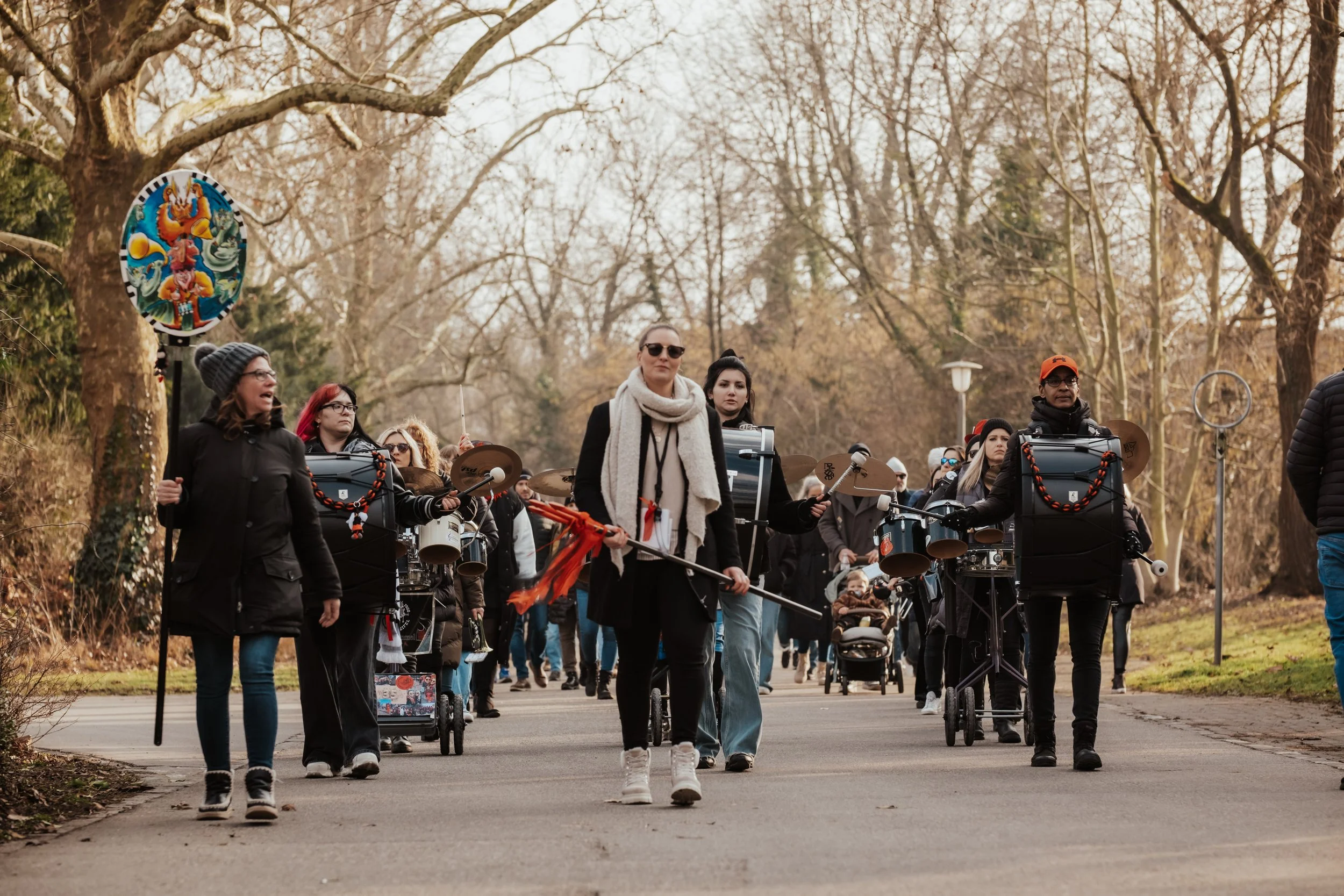 Gruppe von Menschen, die an einem Spaziergang im Park teilnehmen, mit einigen spielenden Musikern, im Hintergrund Bäume ohne Blätter, winterliche Atmosphäre.