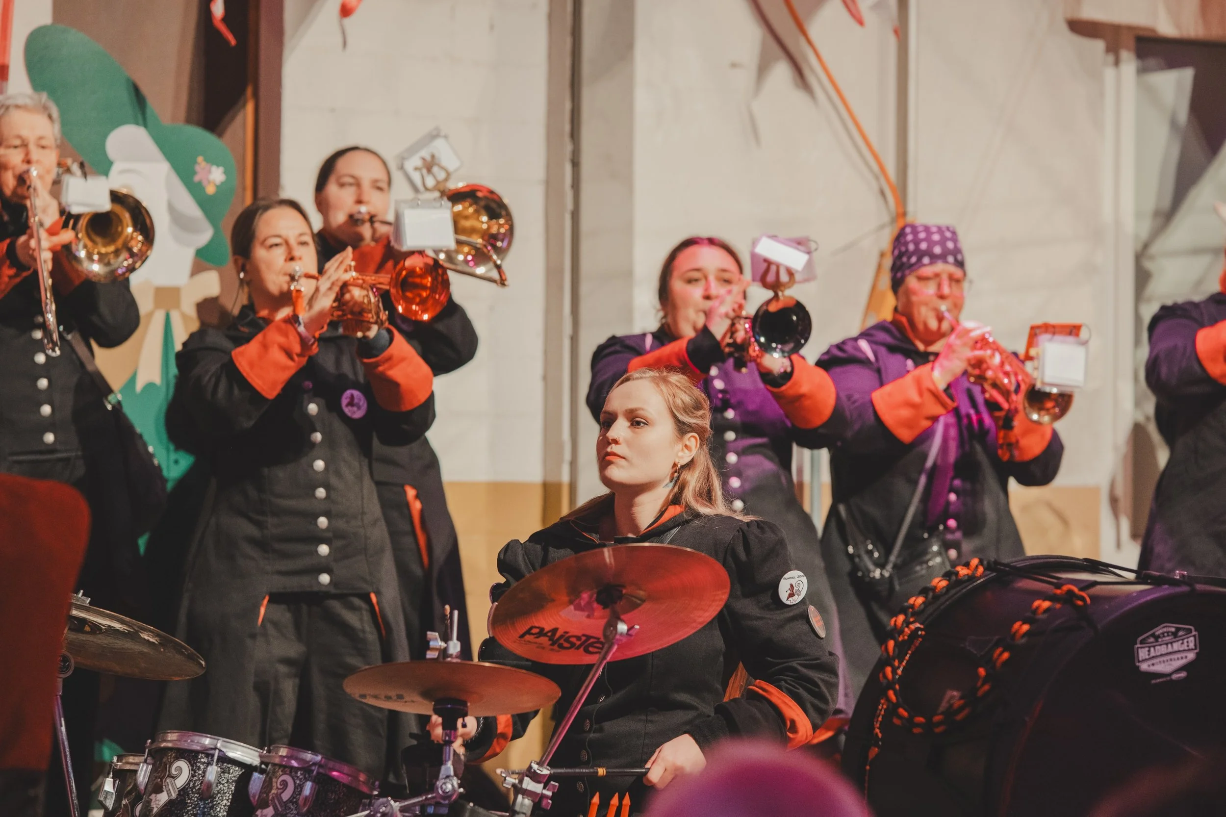 Eine Gruppe von Frauen in schwarzer Kleidung mit roten Akzenten spielt Blasinstrumente und Schlagzeug bei einem Konzert oder einer Feier.