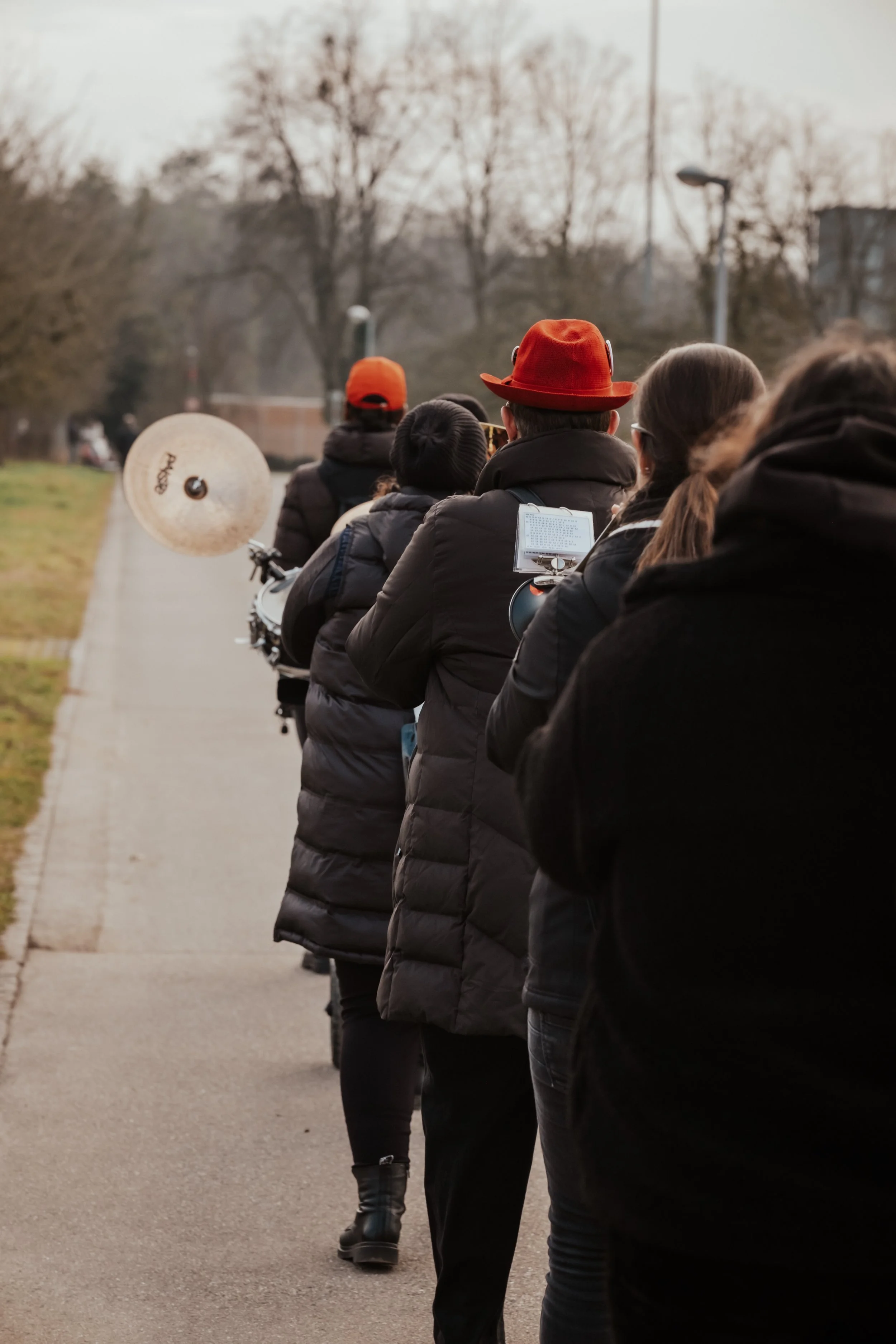 Menschen in Winterjacken stehen in einer Warteschlange auf einem Weg, hinter ihnen ein Musiker mit rotem Hut spielt auf einem Schlagzeug, im Hintergrund sind Bäume ohne Blätter zu sehen.