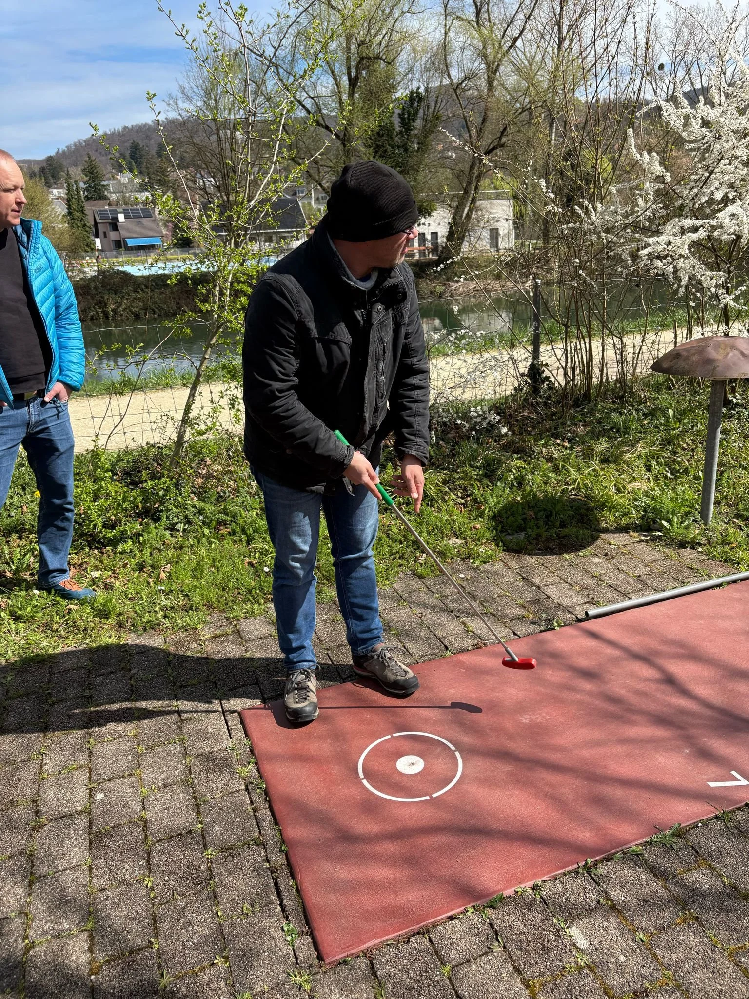 Zwei Männer spielen Minigolf im Freien. Einer schlägt auf den Ball, der auf einer roten Matte liegt, während der andere zuschaut. Es ist Frühling mit blühenden Bäumen im Hintergrund.