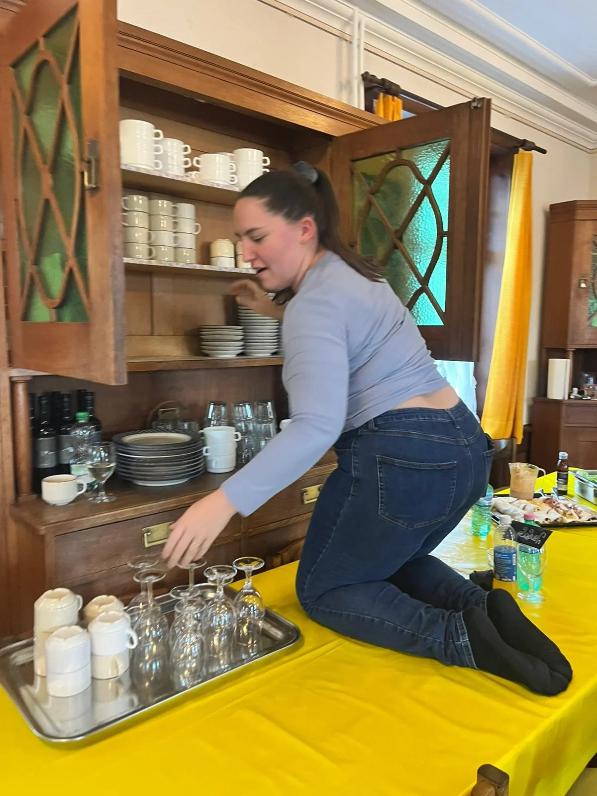 Eine junge Frau hebt im Halbknien auf einem gelben Tisch Geschirr und Gläser auf, während sie in einer Holzkommode mit offenen Schranktüren nach Geschirr greift.