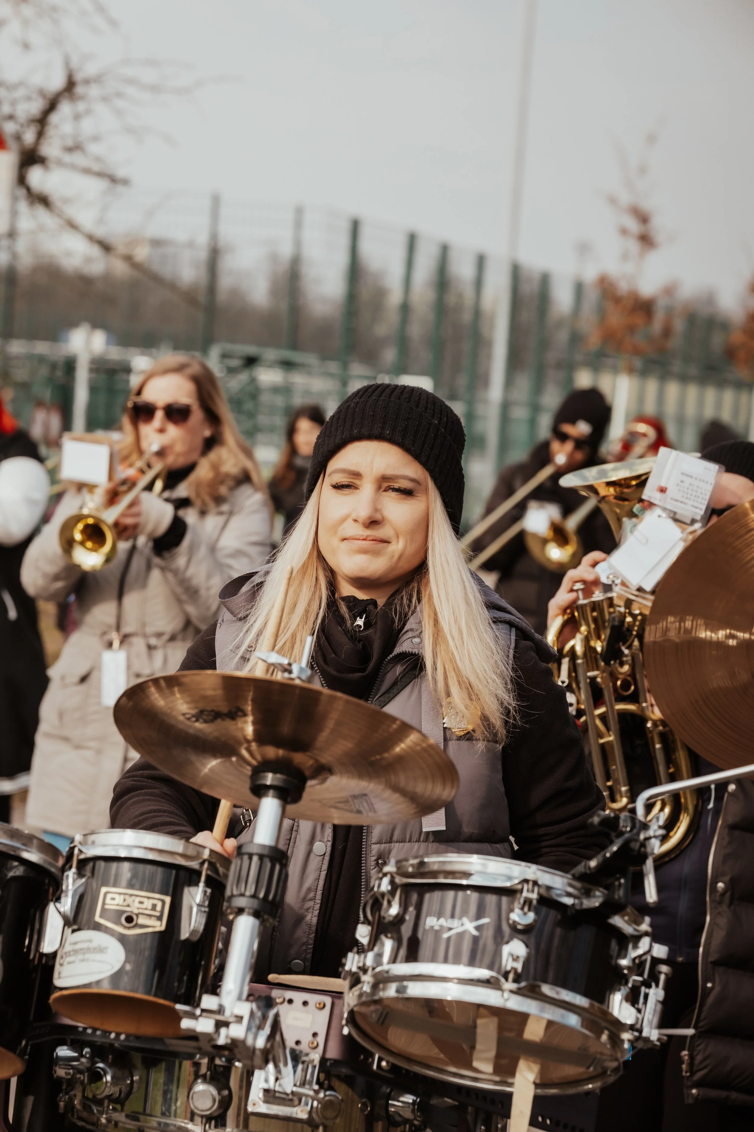 Eine Gruppe von Musikern, darunter eine Frau, die Schlagzeug spielt, während andere Musiker Blasinstrumente spielen, im Freien bei kühlem Wetter, mit kahlen Bäumen im Hintergrund.