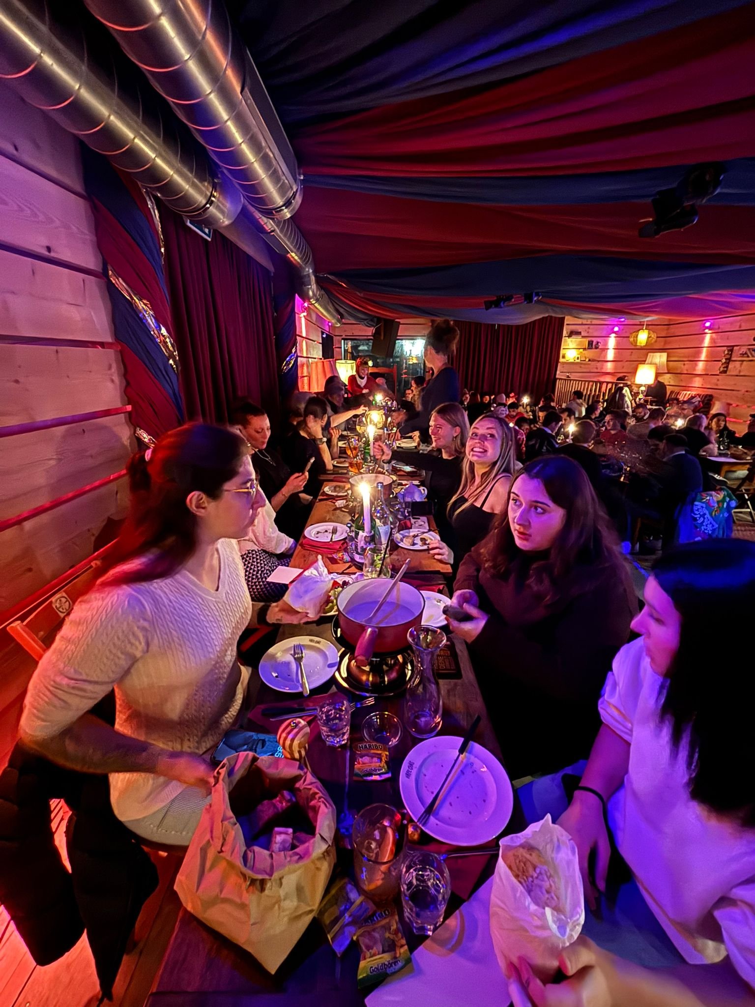 Menschen sitzen an einem langen Tisch in einer gemütlichen Bar oder einem Restaurant, umgeben von bunten, weichen Lichterketten, mit dekorativen Lampen und Stoffbahnen an der Decke, während sie Getränke und Essen genießen.