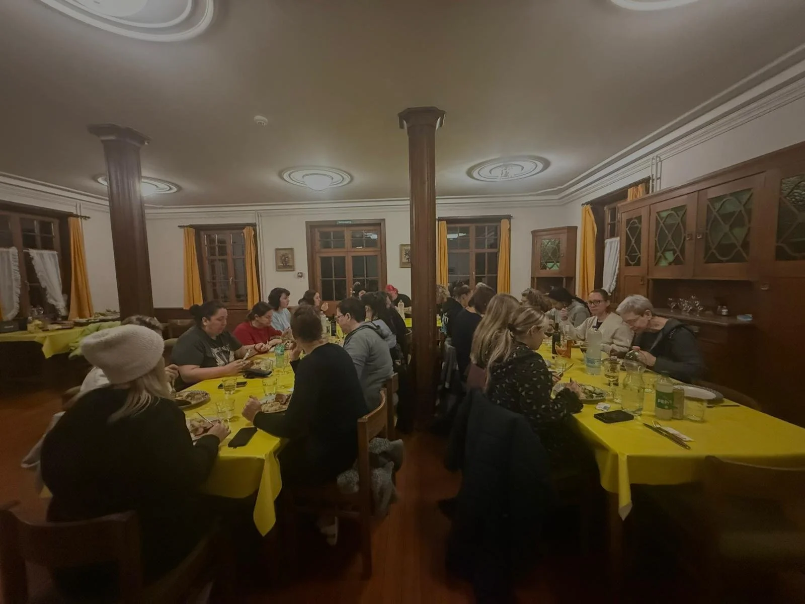 Menschen sitzen an einem langen Tisch mit gelben Tischtüchern bei einem Abendessen in einem rustikalen, holzverzierten Raum.