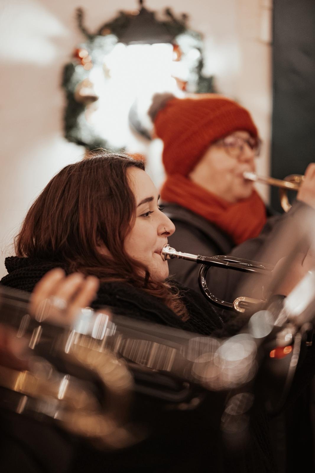 Zwei Personen spielen in einer weihnachtlich dekorierten Umgebung Querflöte. Die Person im Vordergrund hat braune Haare und trägt einen schwarzen Schal. Die Person im Hintergrund trägt eine rote Mütze und einen roten Schal, spielt ebenfalls Querflöte