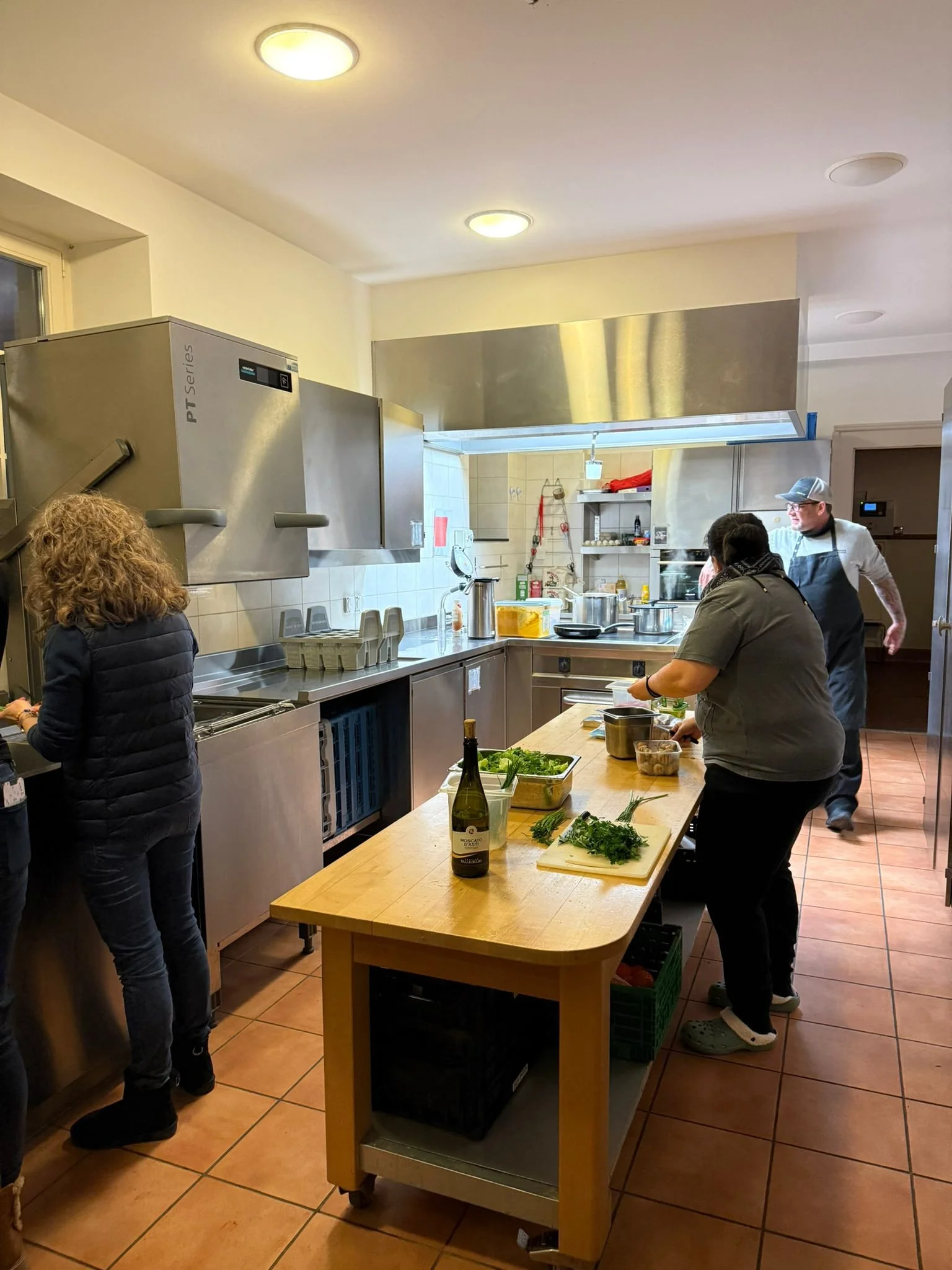 Menschen arbeiten in einer professionellen Küche, mit Küchenschränken, einem Holztisch mit frischen Kräutern, einem Wein, Pfannen und Kochutensilien.