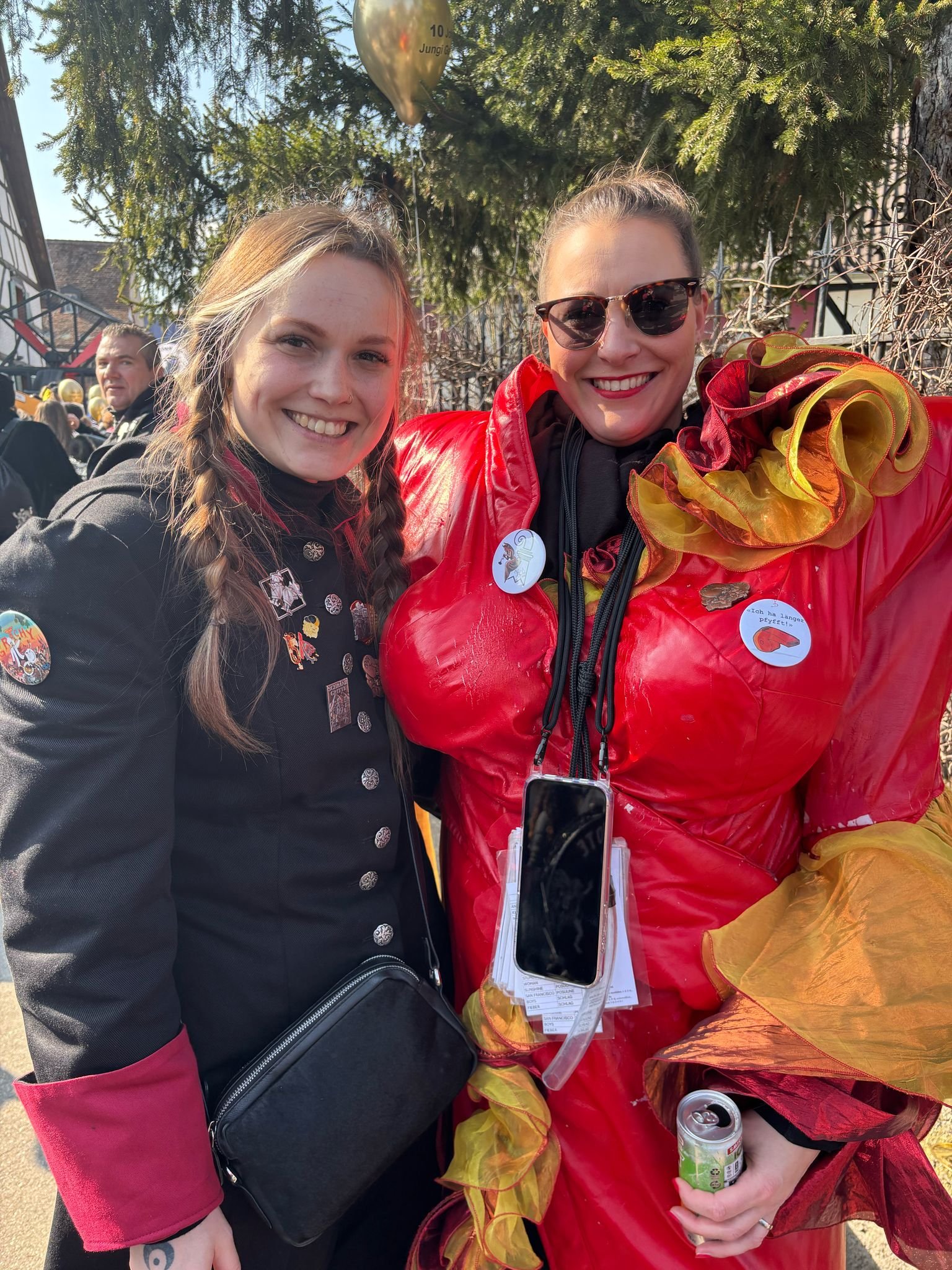 Zwei lebhafte Frauen auf einer Feier, eine trägt ein rotes und gelbes Kostüm mit Rüschen, die andere trägt ein schwarzes Kleid mit Pins. Sie lachen und posieren zusammen unter einem Baum.