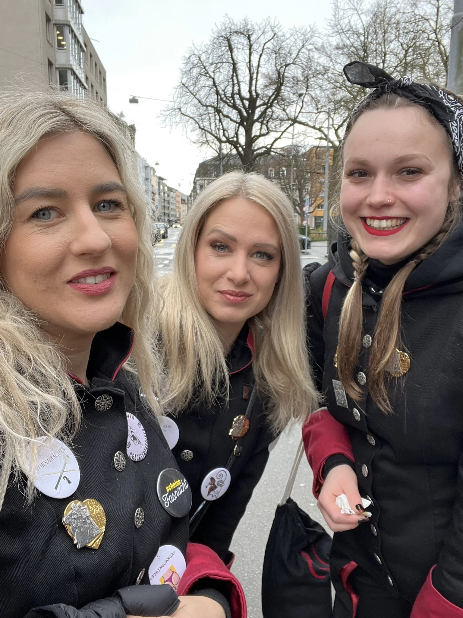 Drei junge Frauen in schwarzer Tracht mit Pins und Button, unterwegs auf einer Straße in einer Stadt, mit Bäumen und Gebäuden im Hintergrund, während des Tages.