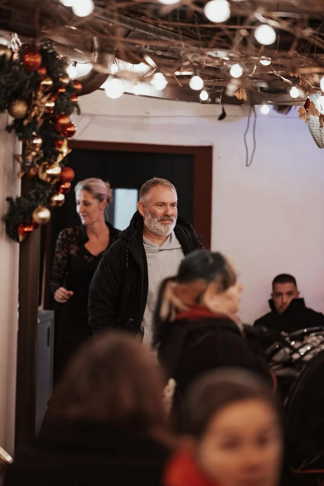 Eine Gruppe von Menschen in einem festlich dekorierten Raum mit Weihnachtsgirlande und Lichterketten, einige sitzen und andere stehen, während sie an einem Weihnachtsfest teilnehmen.