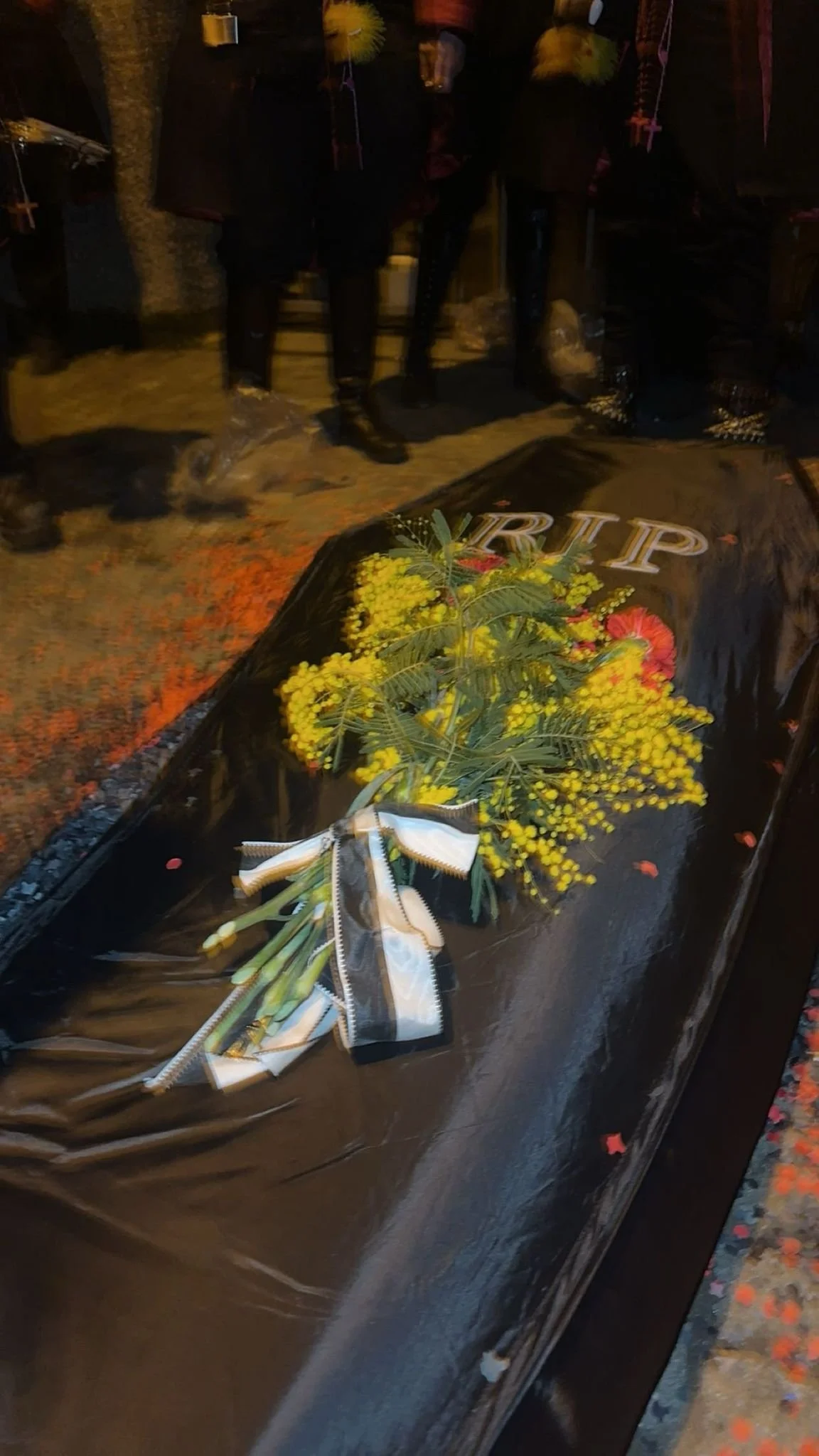 Blumen auf einem schwarzen Grabstein mit dem Wort 'RIP' in goldenen Buchstaben, im Hintergrund Menschen bei einer Gedenkveranstaltung