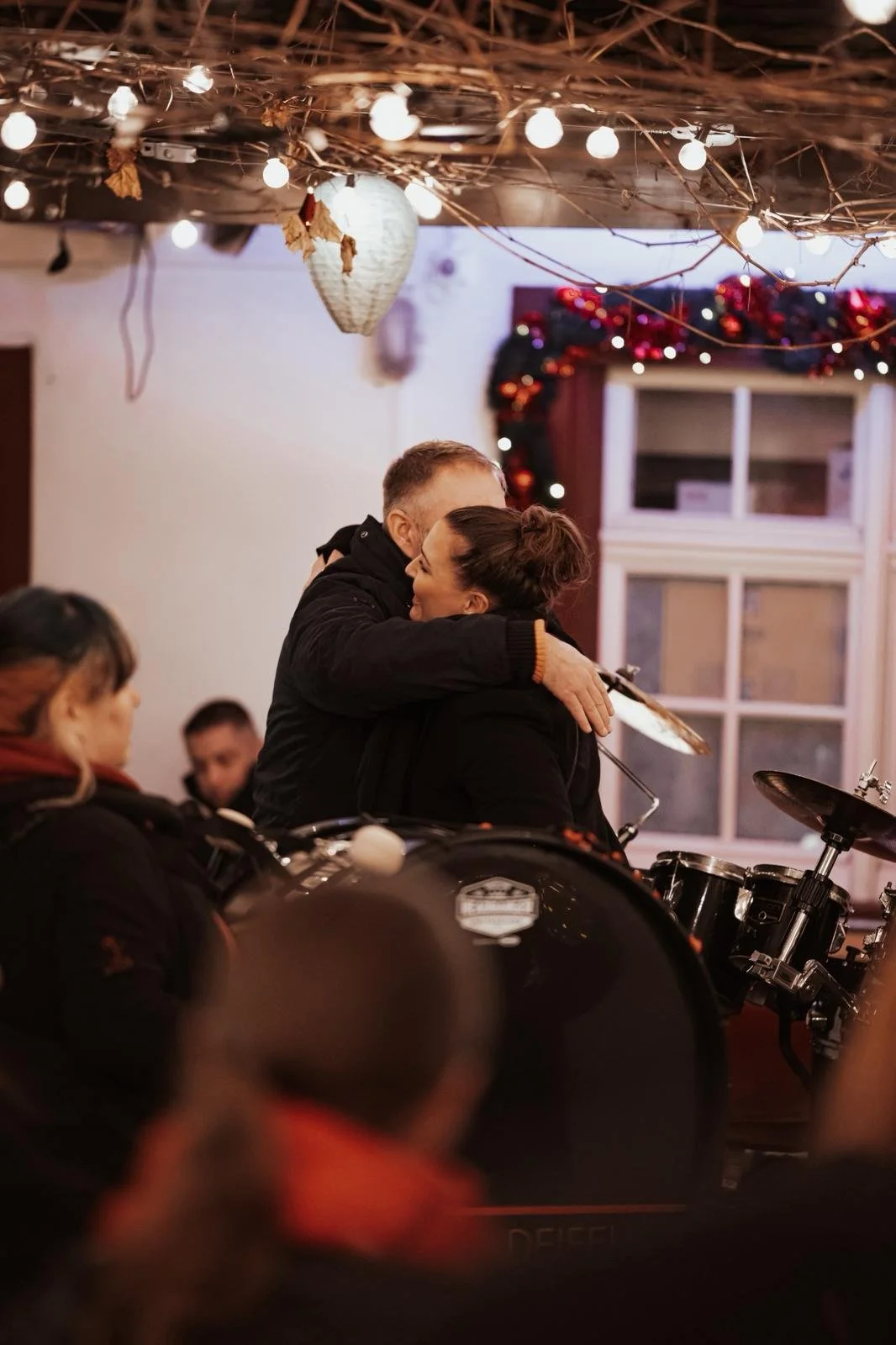 Zwei Menschen umarmen sich in einer festlich dekorierten Umgebung mit Musikinstrumenten, Lichterketten und Weihnachtsdekoration.