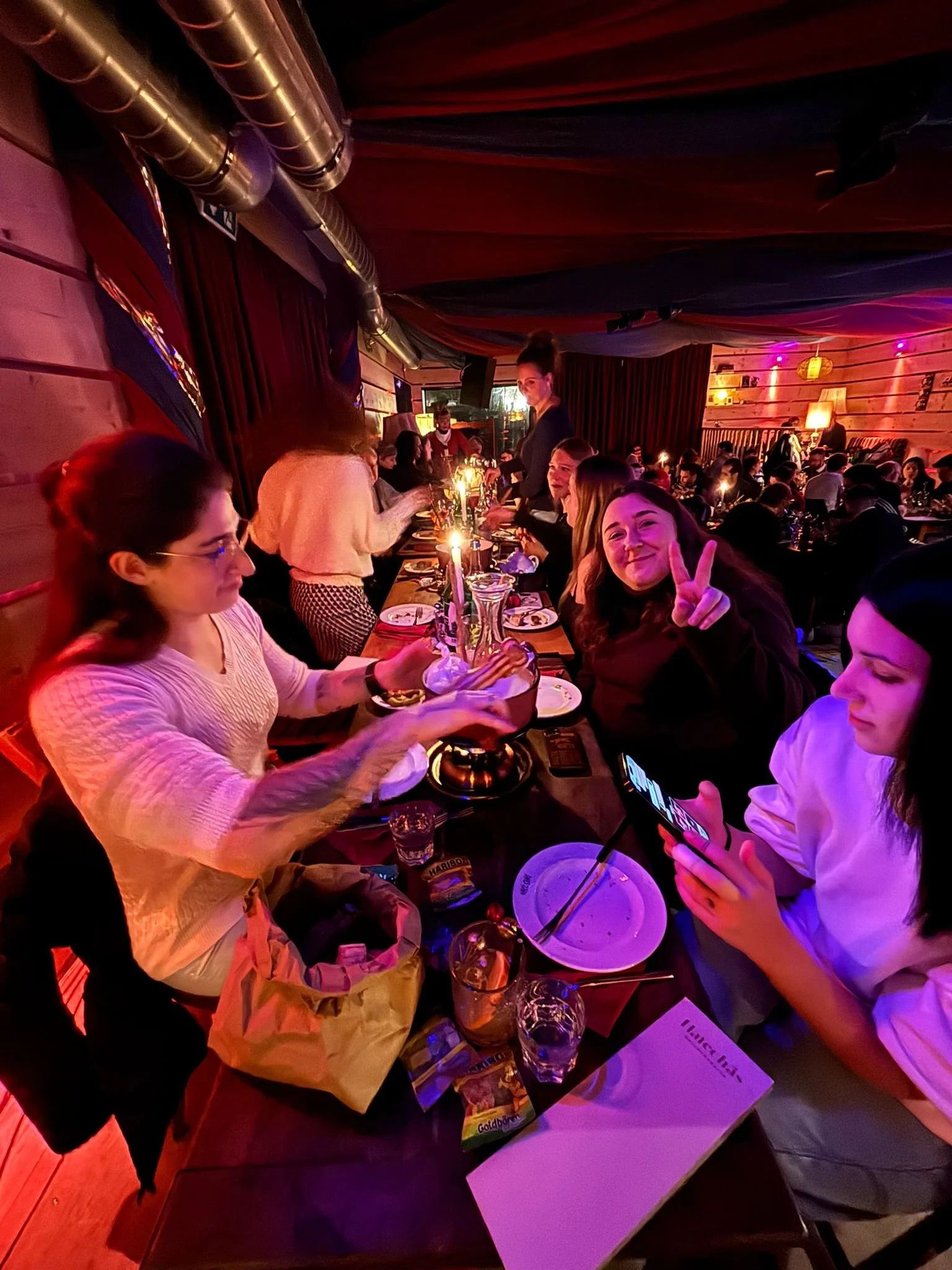 Menschen sitzen an einem langen Holztisch in einer gemütlichen, schummrigen Bar oder Restaurant, trinken und unterhalten sich bei Kerzenlicht.