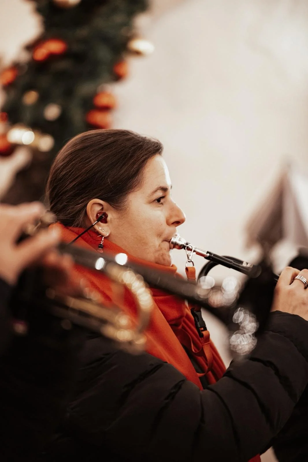 Frau spielt die Trompete, trägt in Ear-Headsets, trägt eine rote Jacke, im Hintergrund ein Weihnachtsbaum mit Ornamenten.