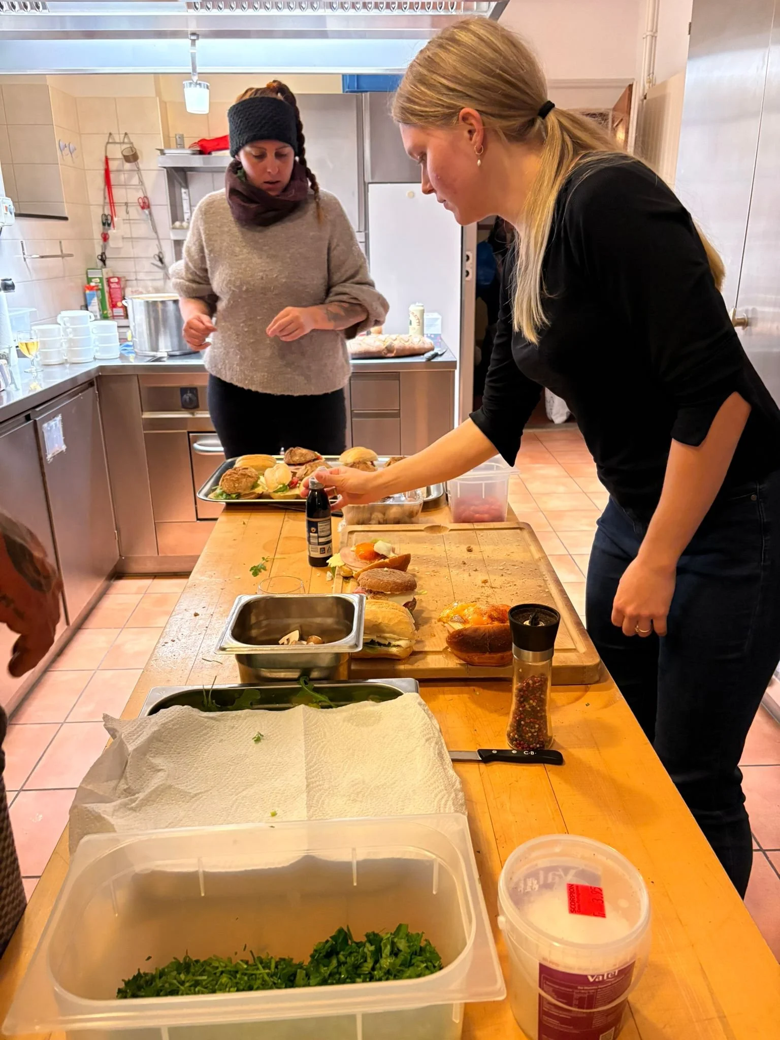 Zwei Frauen bereiten Sandwiches in einer Küche vor, auf einem Tisch sind verschiedene belegte Brötchen, ein Messer, Pfeffer und Salat. Im Hintergrund gibt es Küchenutensilien und einen Ofen.