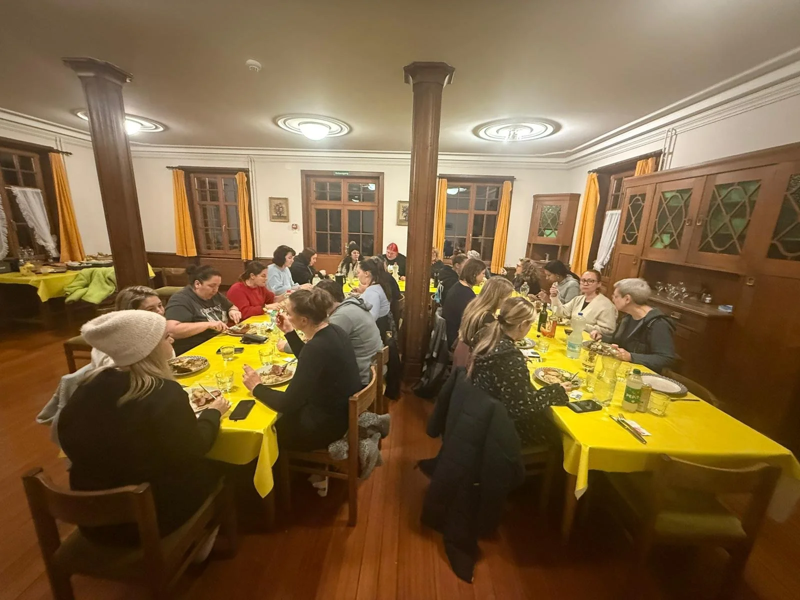 Mehrere Menschen sitzen an zwei Tischen in einem gut beleuchteten Raum, essen und trinken. Die Tische sind mit gelben Tischtüchern bedeckt.