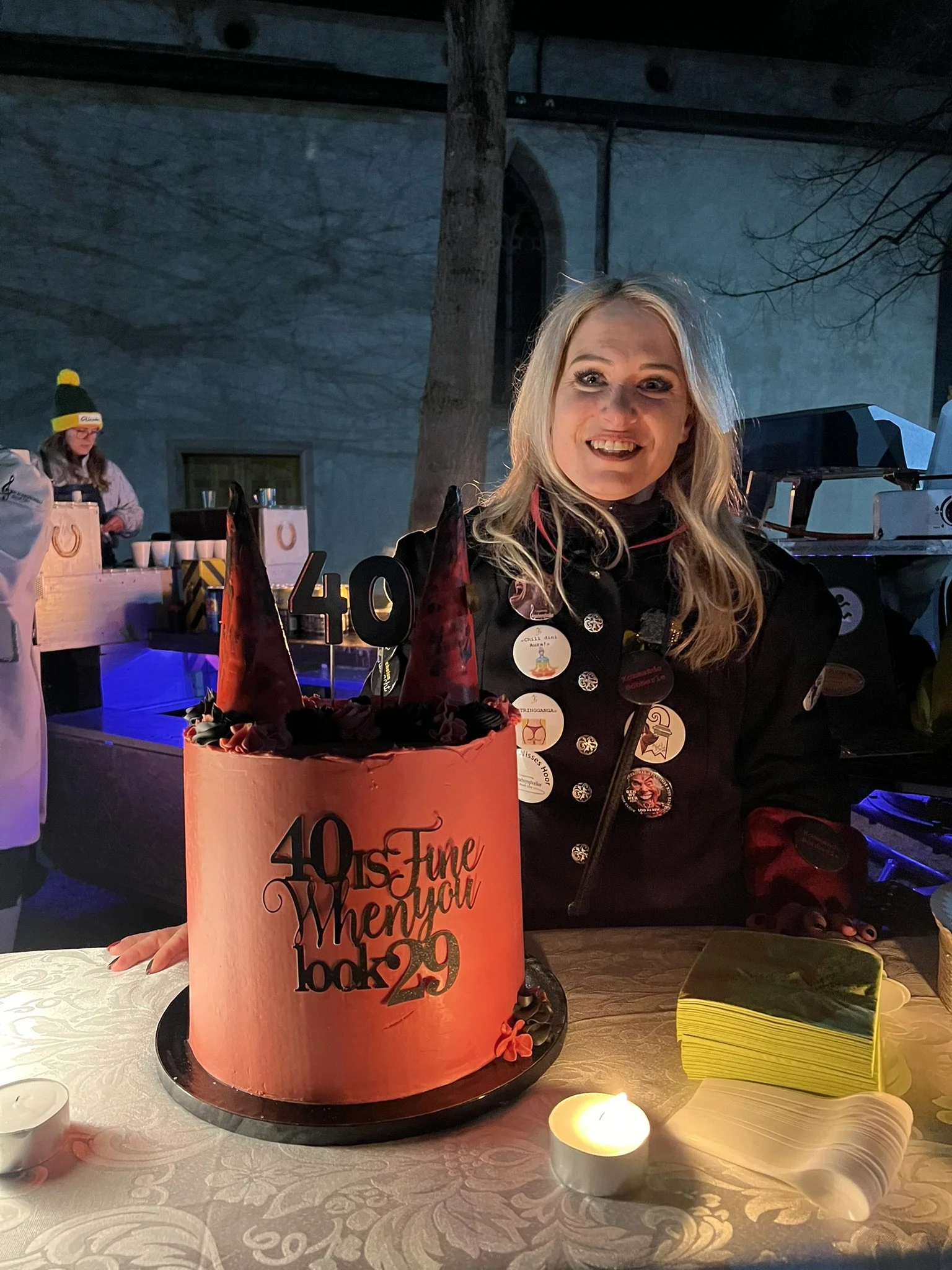 Eine Frau sitzt an einem Tisch mit einer großen Geburtstagstorte, auf der steht '40 is Fine, Many happy Looks'. Die Frau lächelt. Im Hintergrund sind andere Personen und Bäume sichtbar.