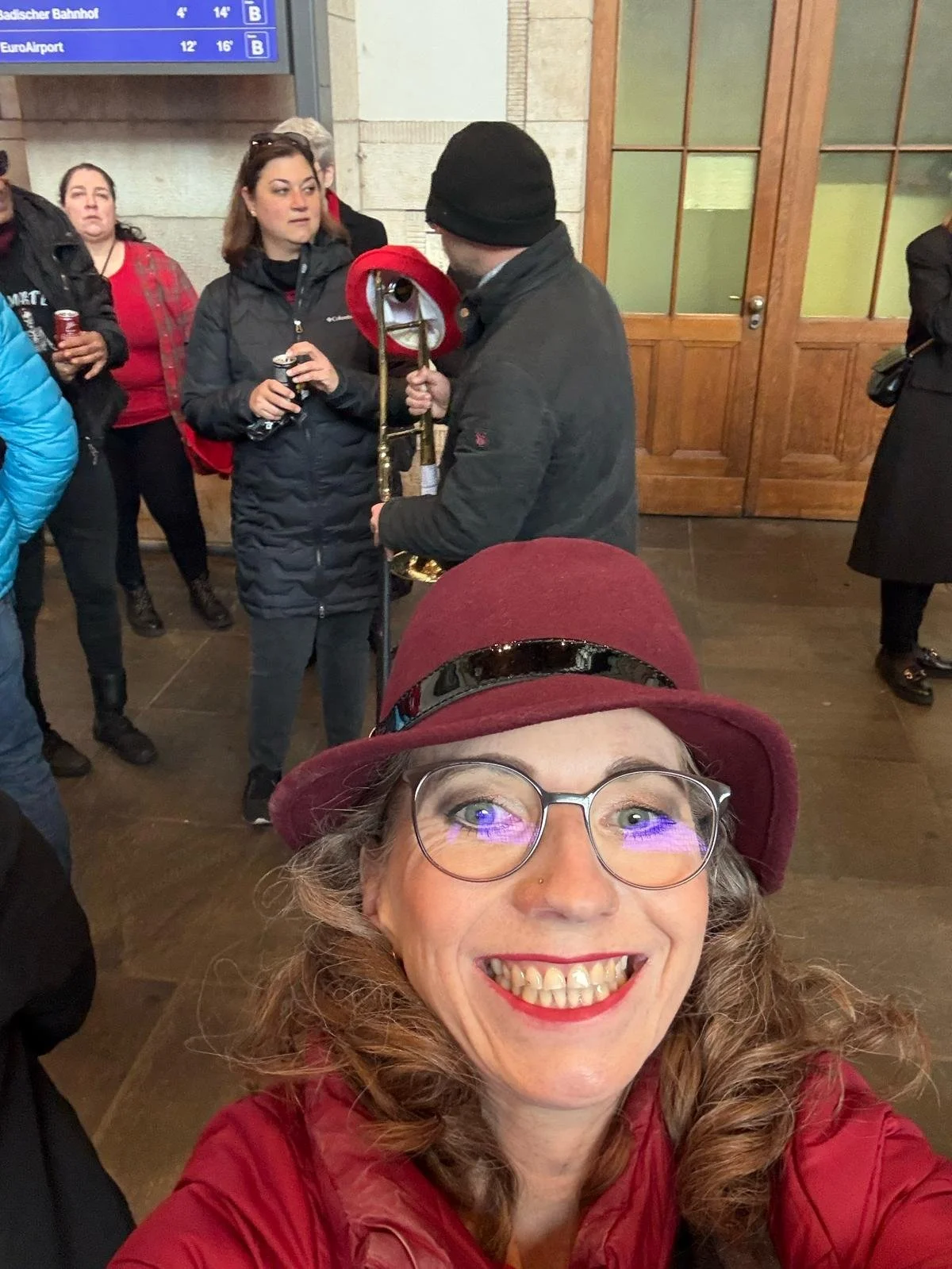 Eine fröhliche Frau trägt eine rote Mütze und eine rote Jacke, trägt Brille und hat lockiges Haar, sie macht ein Selfie in einem Bahnhof mit anderen wartenden Menschen im Hintergrund.