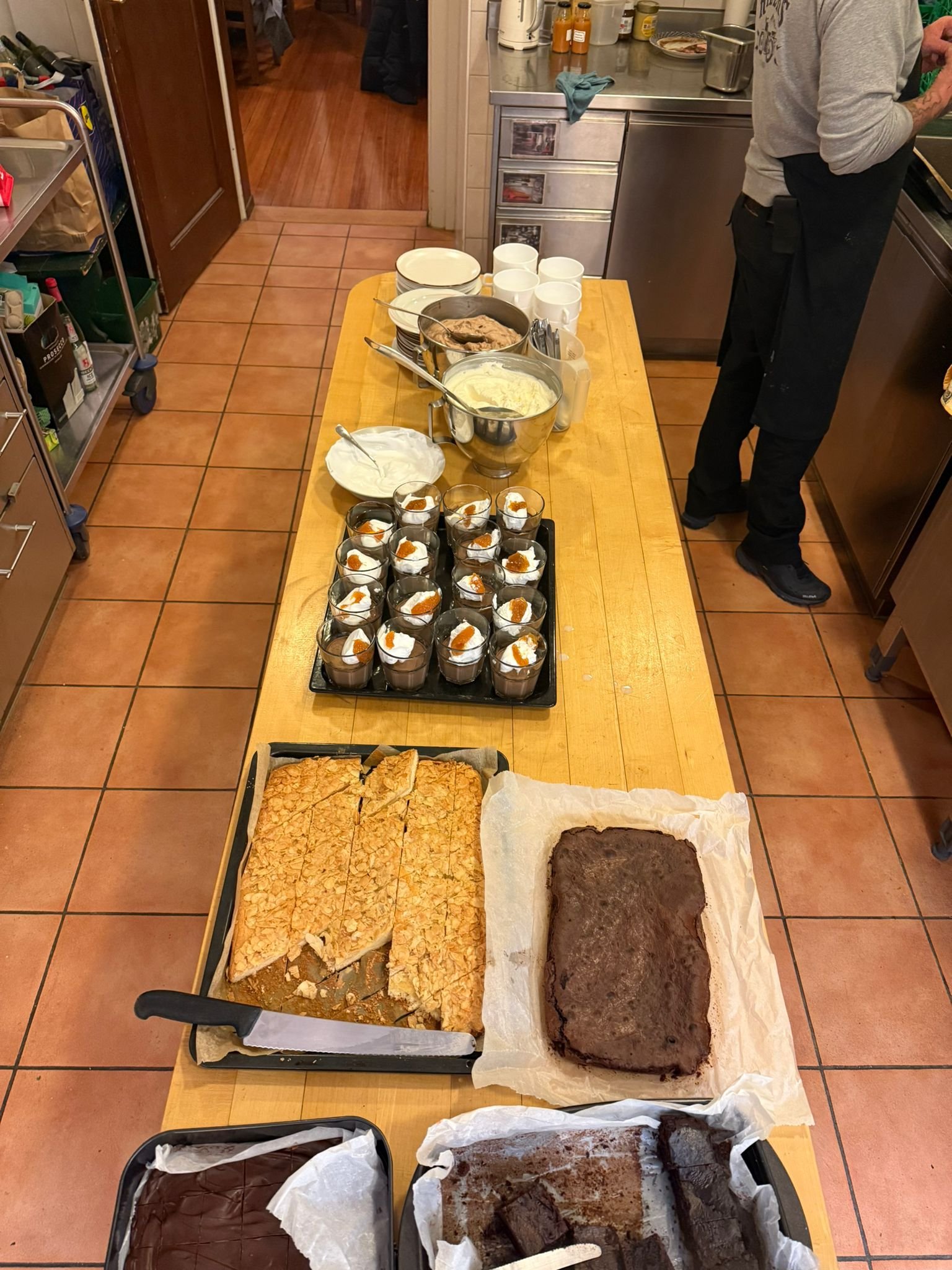 Verschiedene Desserts, Kuchen, Pudding und Drinks auf einem langen Holztisch in einer Küche, mit jemandem im Hintergrund