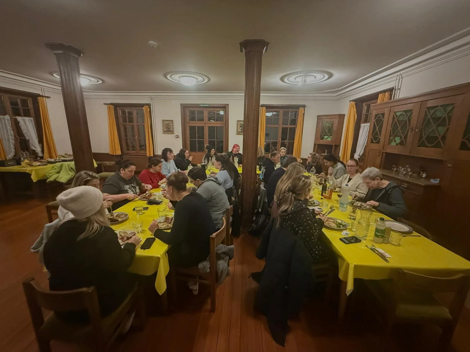 Mehrere Menschen sitzen an zwei langen Tischen mit gelben Tischdecken in einem gut beleuchteten Raum und essen.