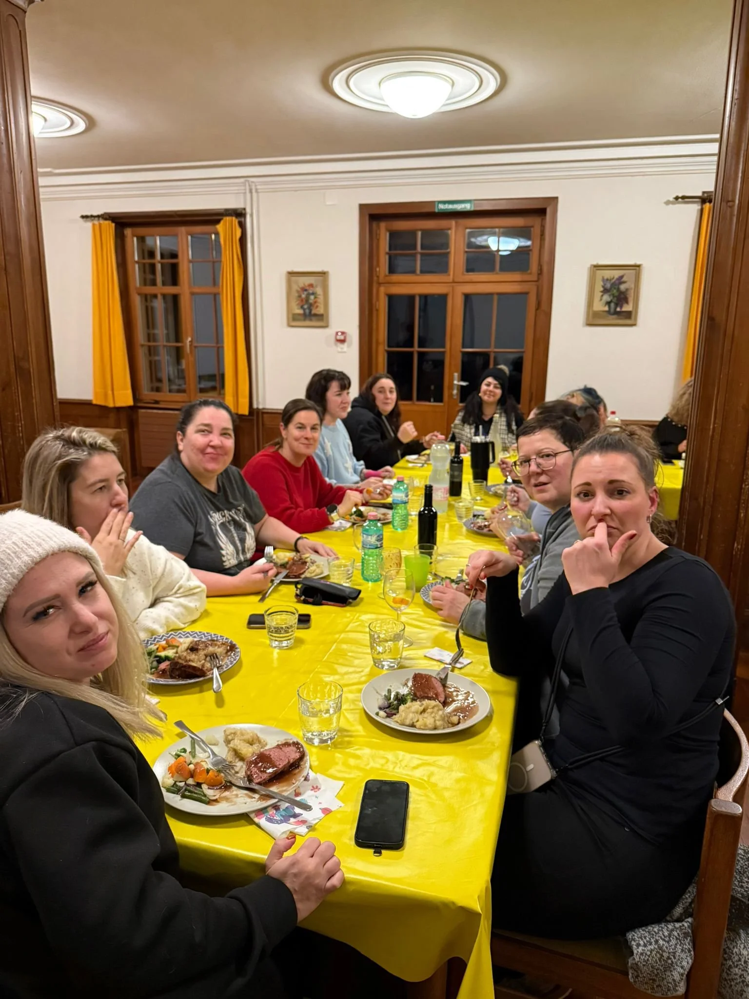 Gruppe von Menschen sitzt an einem langen Tisch mit gelber Tischdecke, isst Mahlzeiten und trinkt Getränke in einem gemütlichen Zimmer mit Holzfenstern und bunten Vorhängen.