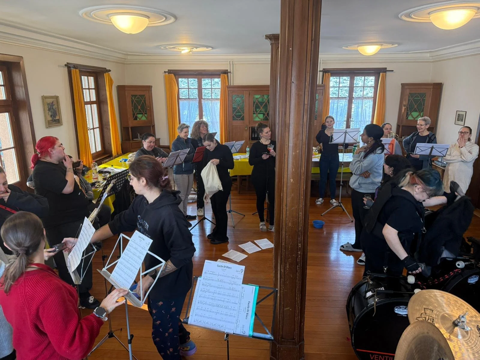 Eine Gruppe von Menschen bereitet sich auf eine musikalische Probe oder ein Konzert vor, mit Notenständern, Musikinstrumenten und einer freundlichen Atmosphäre in einem gut beleuchteten Raum mit großen Fenstern und gelben Vorhängen.