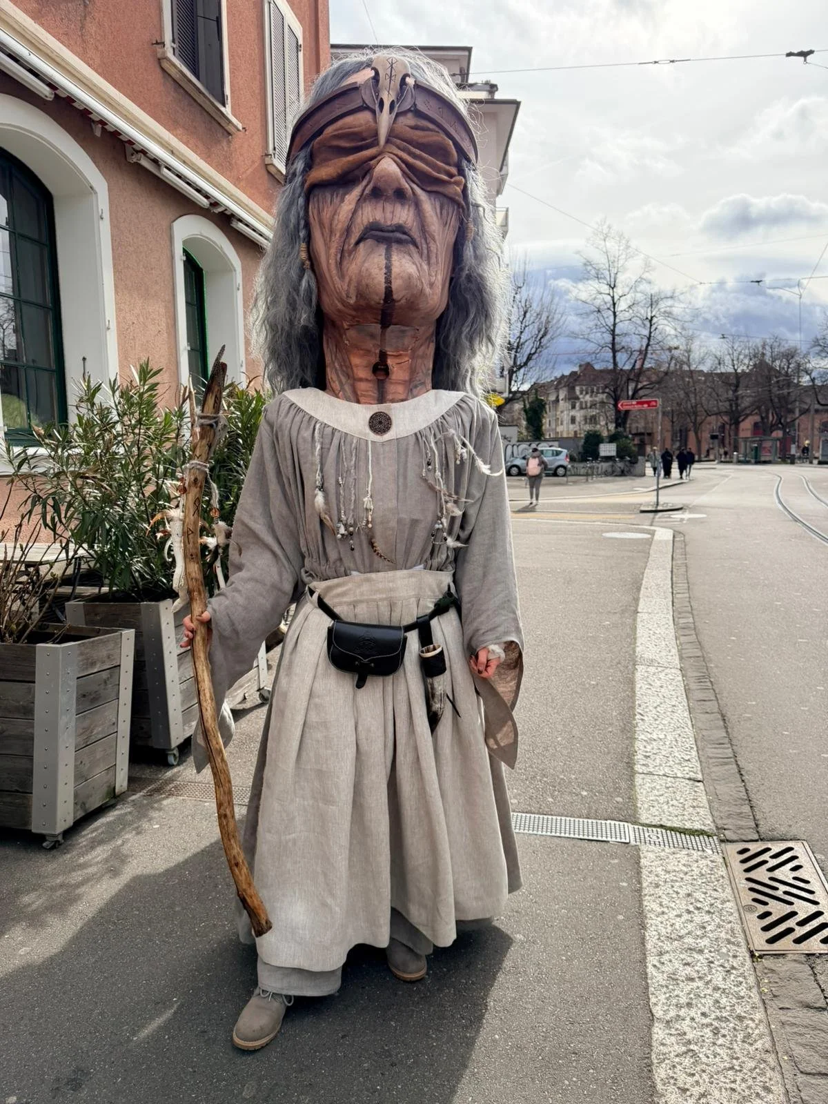 Person in Kostüm mit großer, künstlerisch gestalteter Maske, die wie eine alte, groteske Figur aussieht, auf einer städtischen Straße.