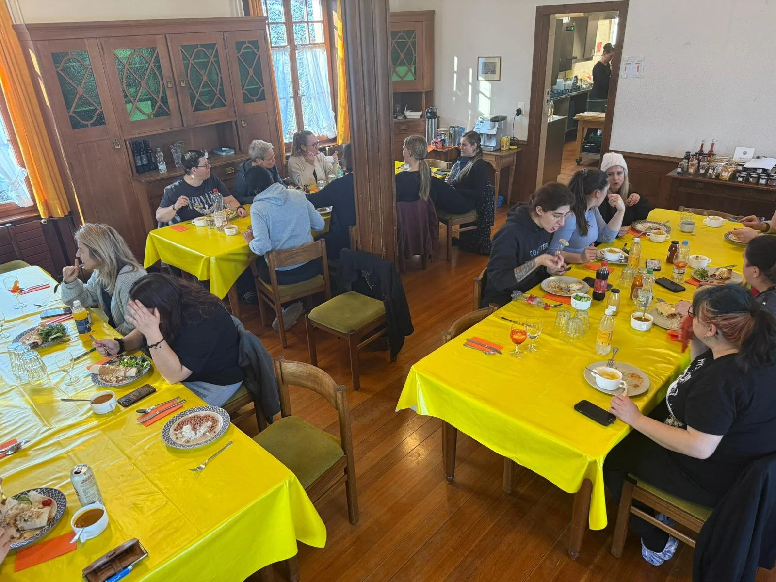 Gruppentisch mit Menschen beim Essen in einem rustikalen Raum mit Holzwänden und gelben Tischdecken.
